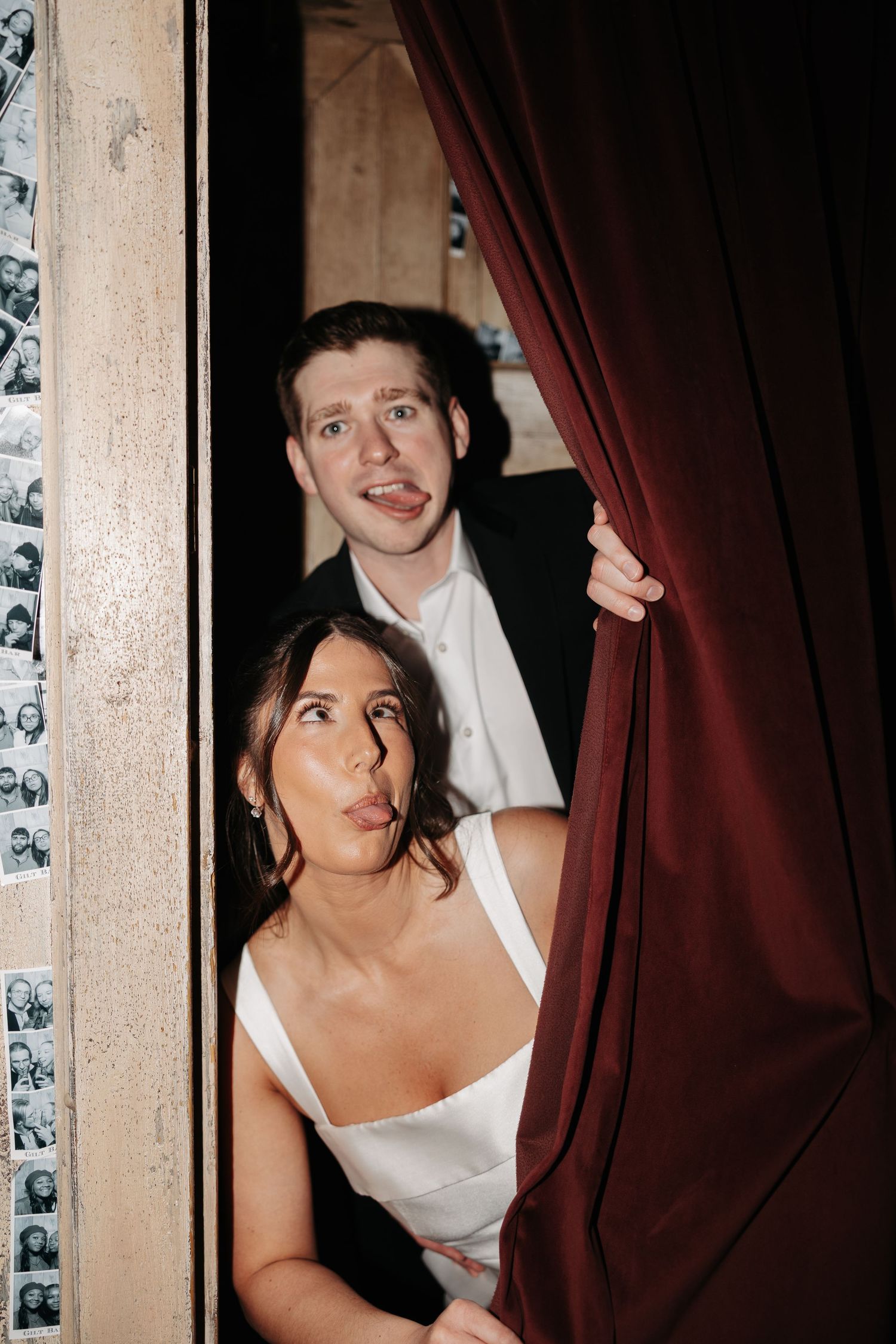 A couple shares a playful moment behind a red curtain in a vintage-style photo booth setting.