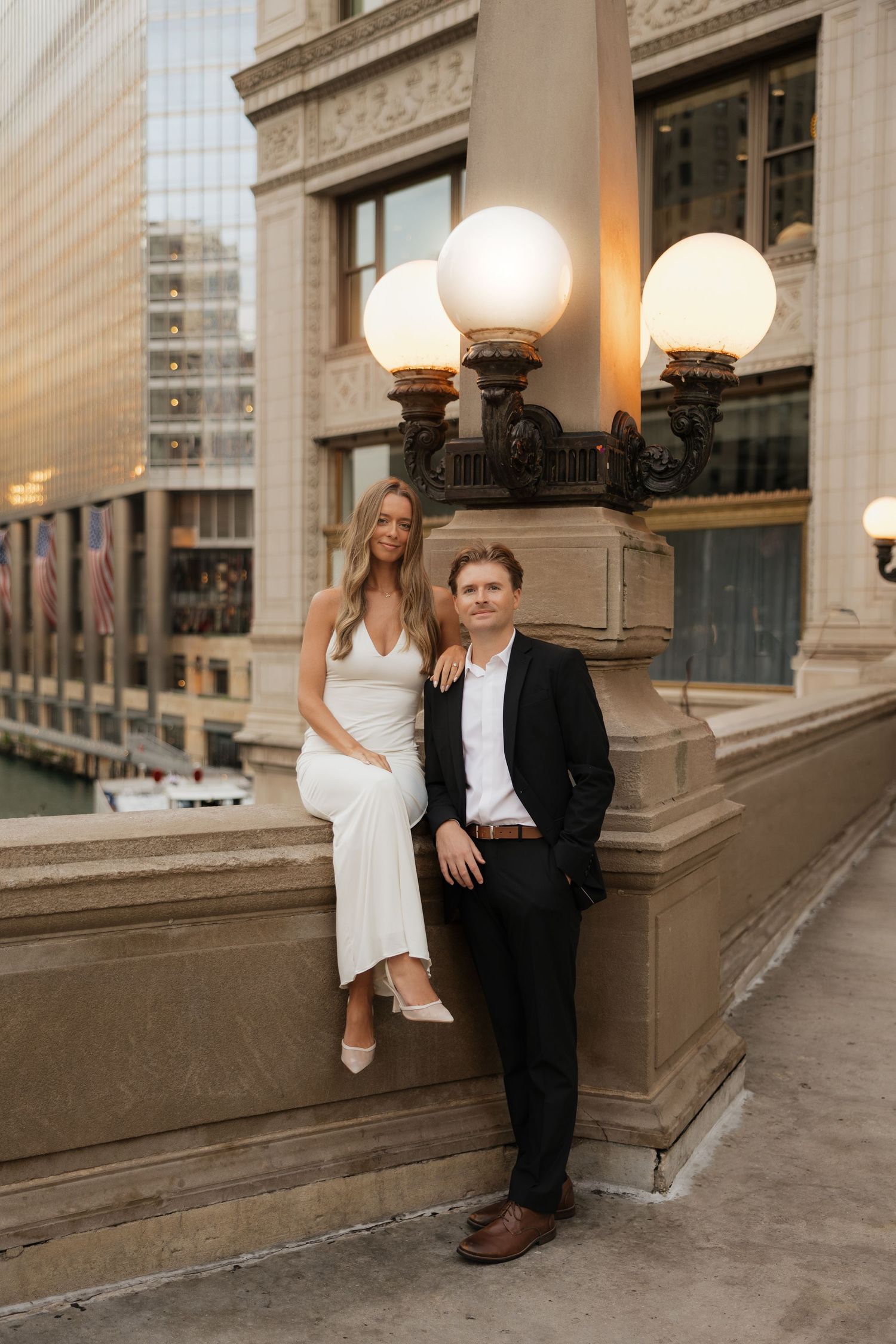 Couple posing by ornate lamp post on Chicago Riverwalk, woman in white dress seated, man in black suit standing.