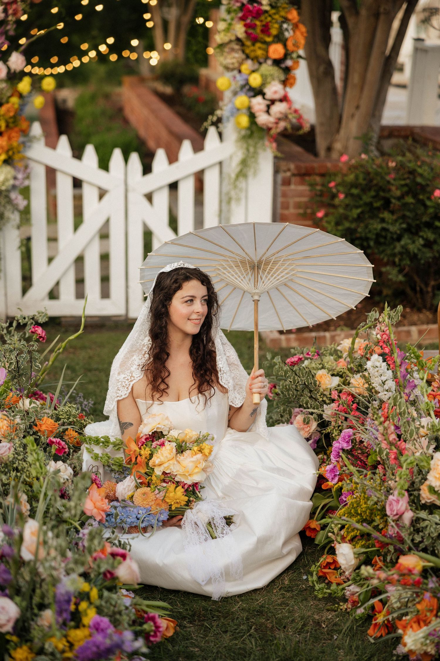 Bride in white gown sits among colorful flowers holding parasol and bouquet near white picket fence.