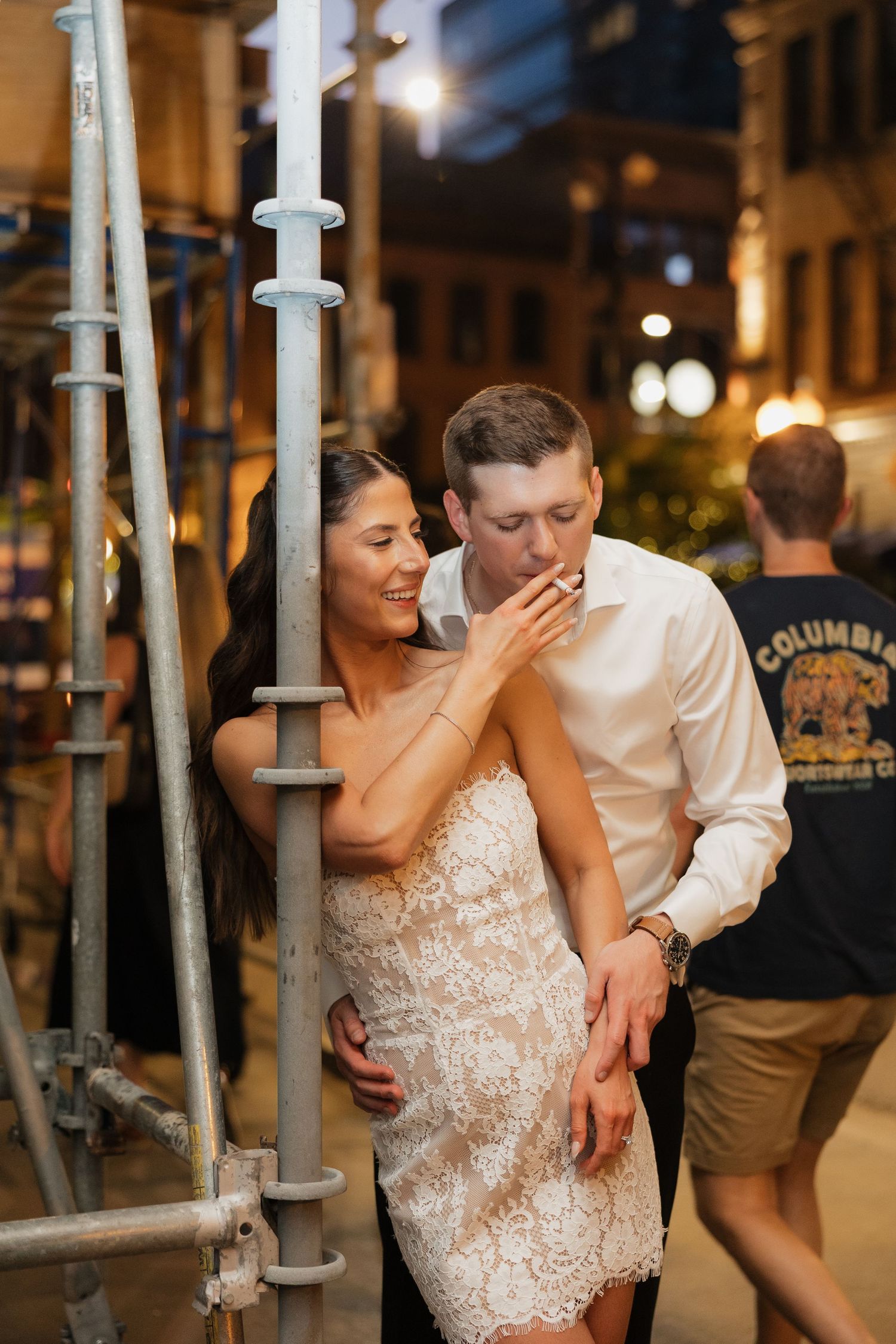 Couple in wedding attire sharing a romantic moment near scaffolding on a city street at night.