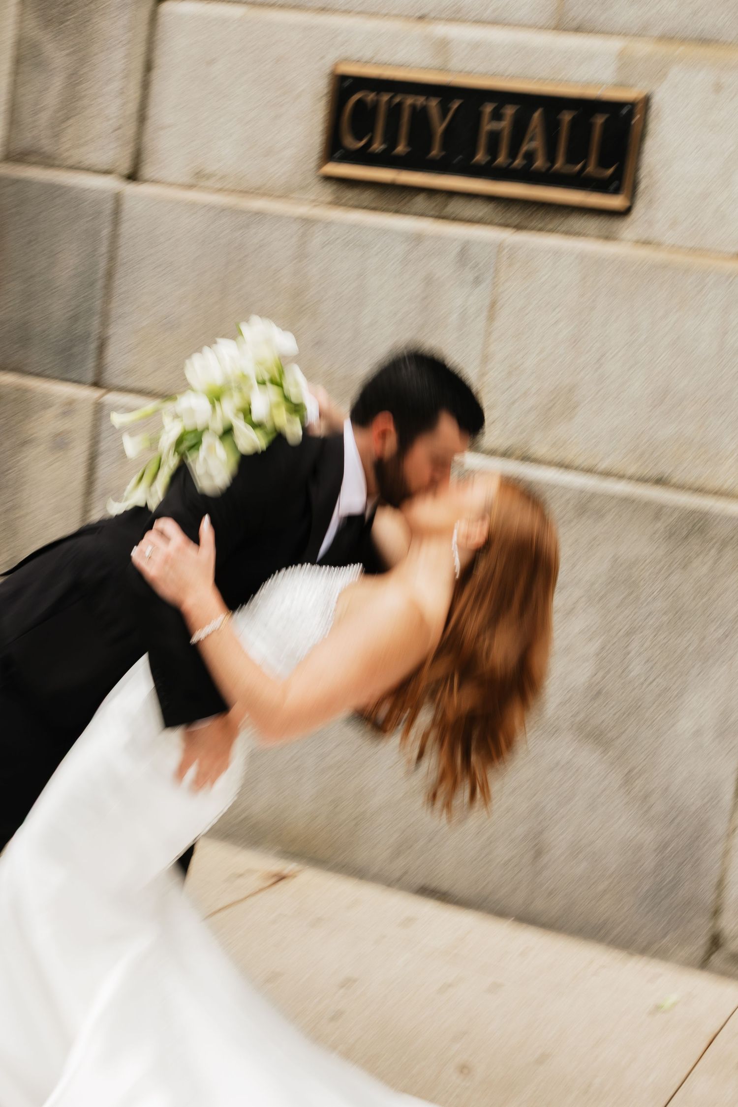 Bride and groom share a romantic dip kiss in front of a City Hall stone wall, bride holding white tulip bouquet.