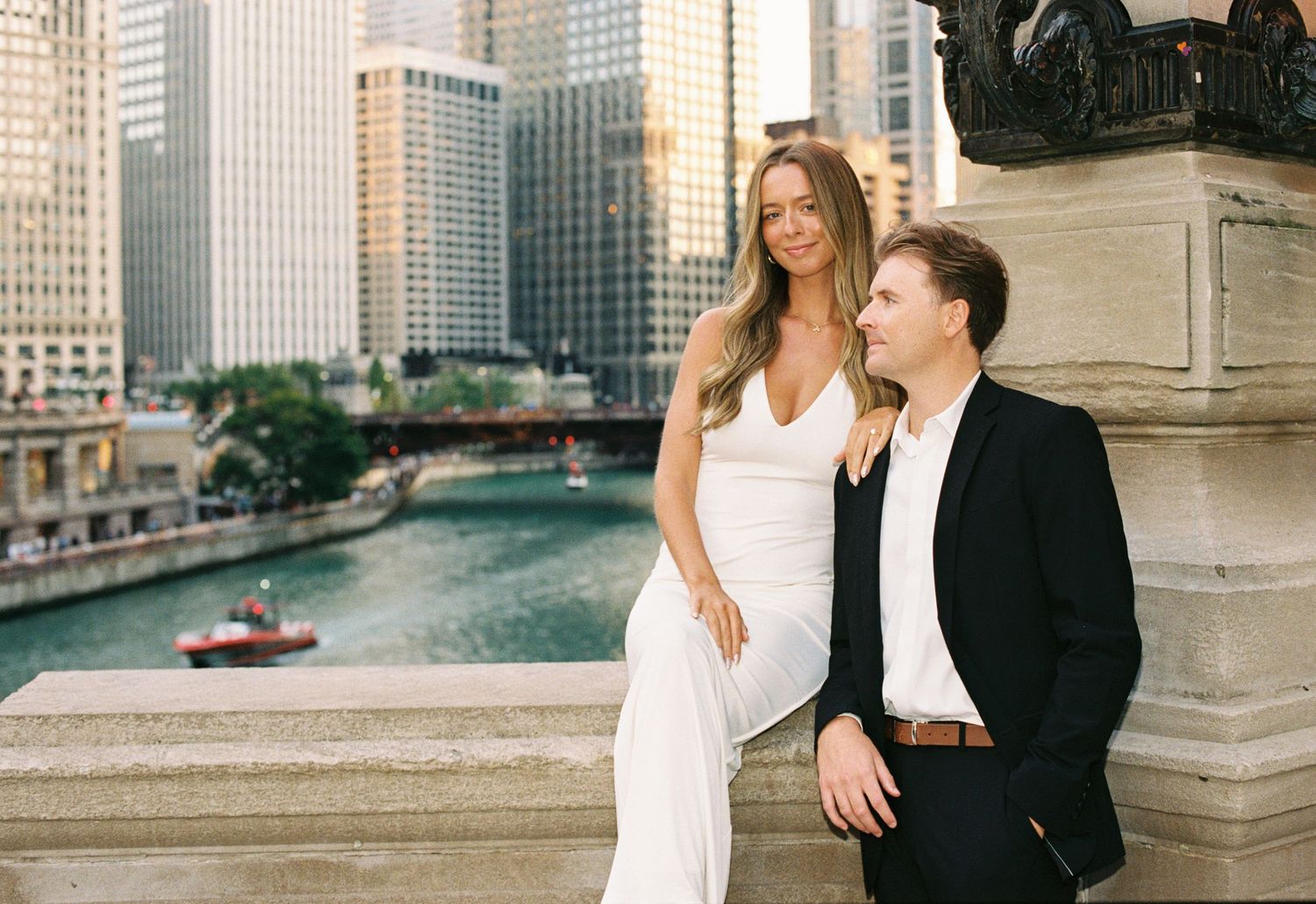 Couple posing by Chicago River with city skyline backdrop, woman in white dress, man in black suit.
