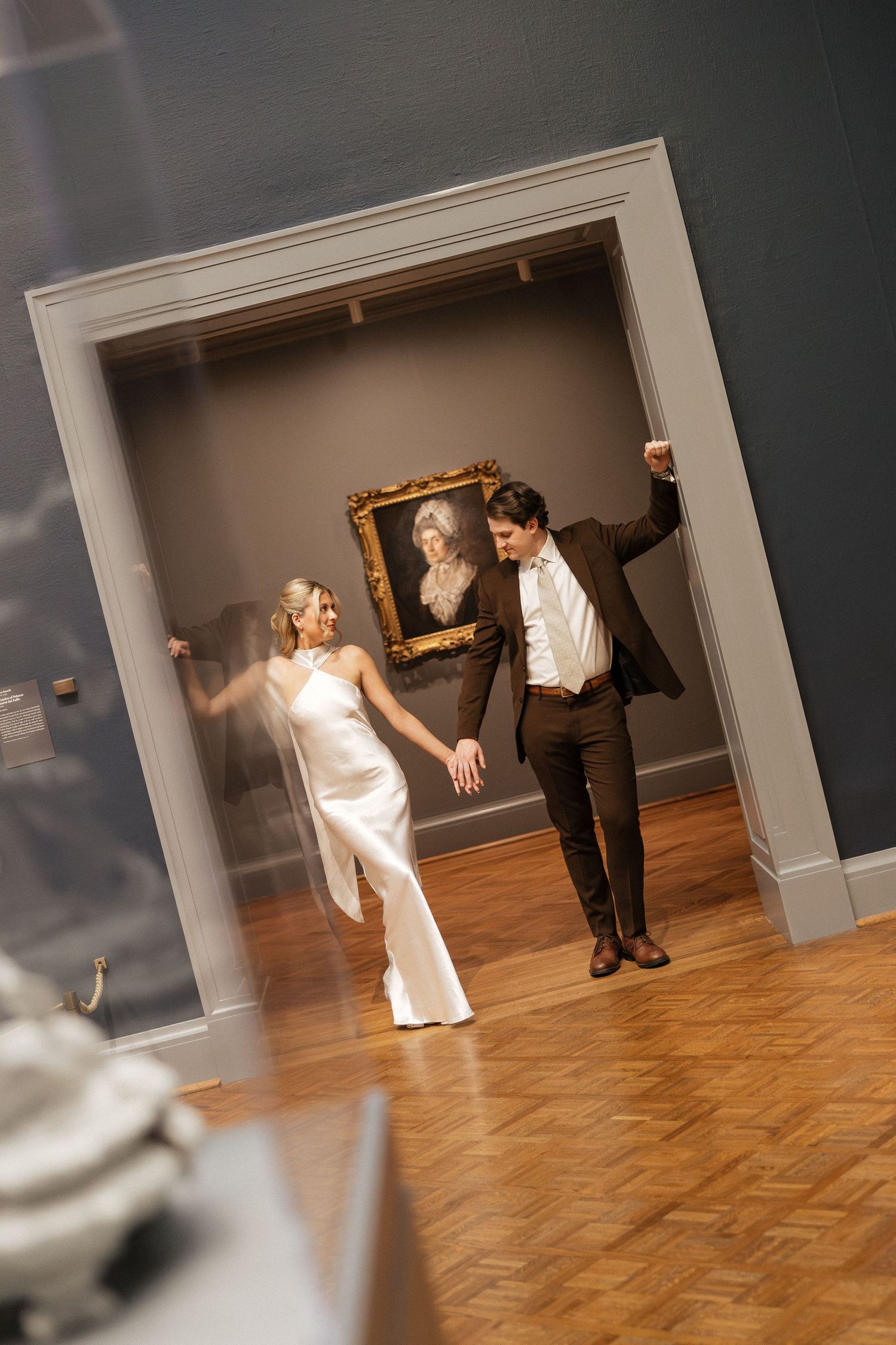 Bride and groom dancing joyfully in an art museum gallery with a classic portrait painting in the background.