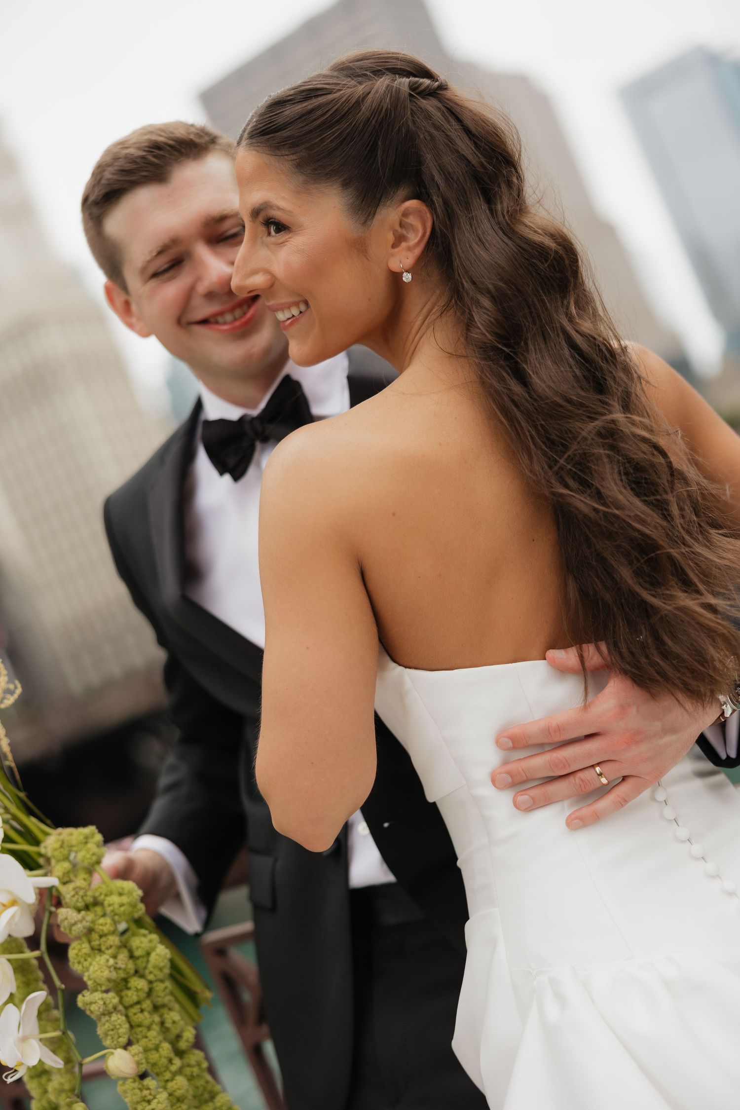 A groom in a tuxedo embraces his bride in a white strapless gown on a rooftop with city views.