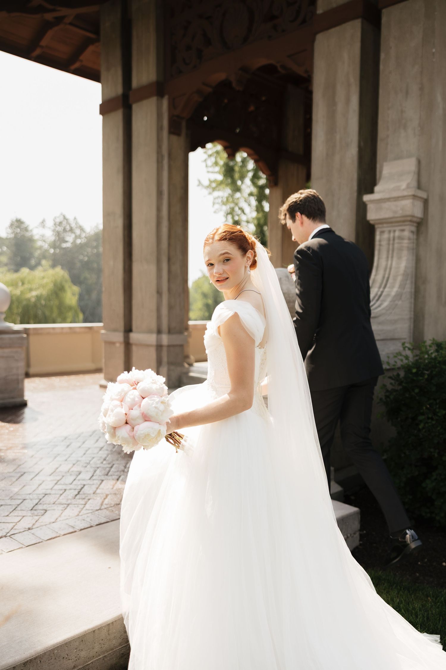 Smiling bride in white gown holding pink bouquet with groom in black tuxedo at elegant stone venue.