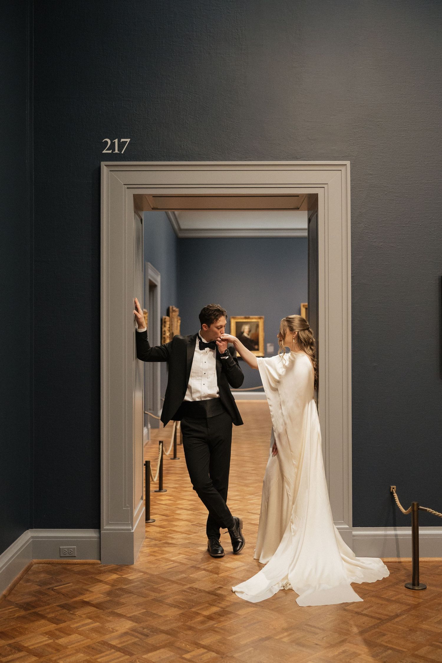 A couple in wedding attire playfully poses in a museum gallery with hardwood floors and white doorways.