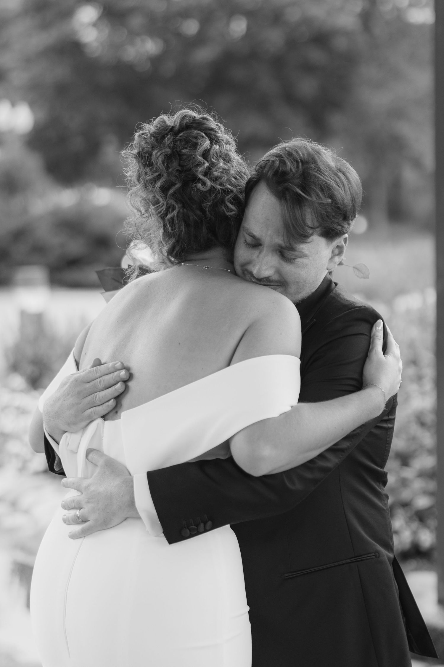 Black and white photo of a bride in an off-the-shoulder gown embracing a groom in a dark suit during an emotional outdoor wedding moment.