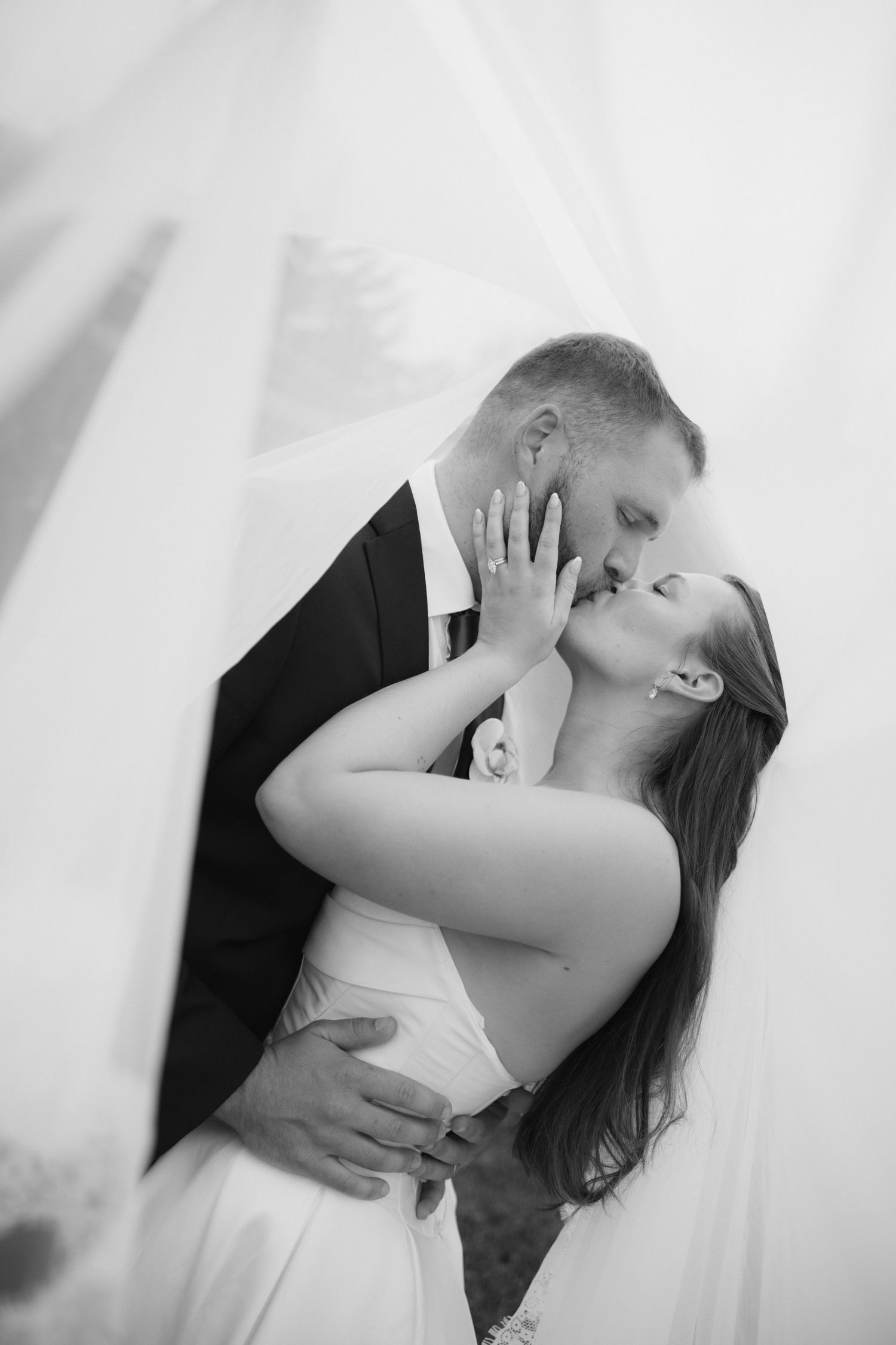 Black and white wedding photo of a bride and groom kissing tenderly as a sheer veil drapes around them, her hand touching his face.