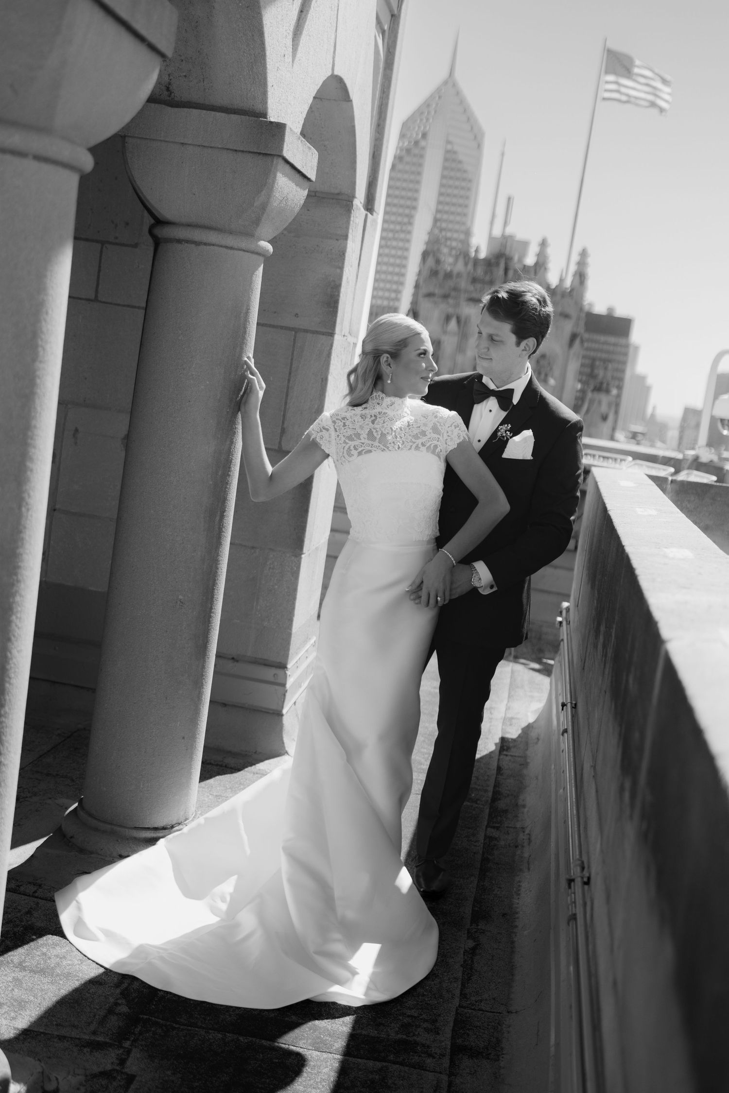 Black and white photo of a bride in a lace-sleeved gown and a groom in a tuxedo embracing on a sunlit city rooftop terrace.