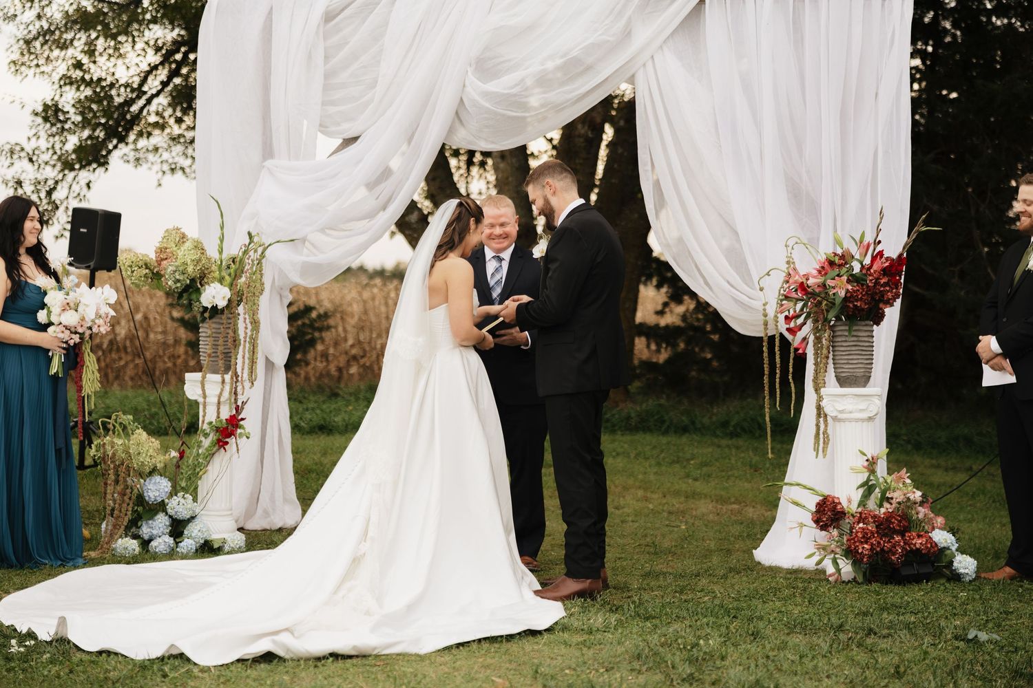 Bride in a long-train white gown and groom face the officiant under draped fabric and floral arrangements at an outdoor wedding.