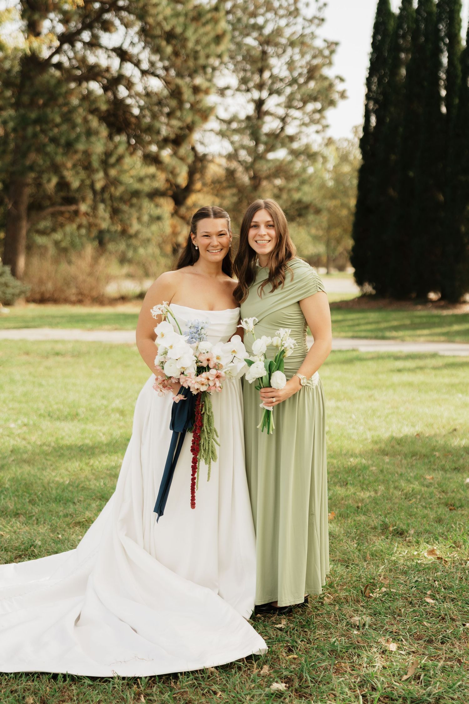 Bride in a strapless white wedding gown and bridesmaid in a sage green dress hold white floral bouquets while posing on a grassy lawn.
