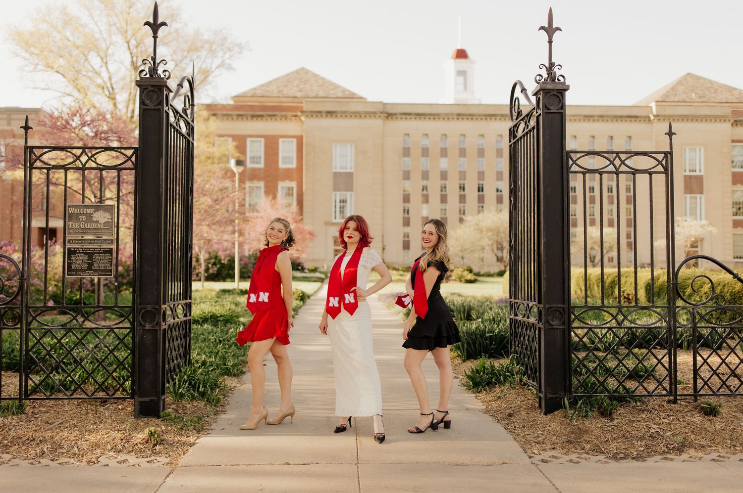 Three friends in red dresses pose together in a series of photos on a college campus with ornate gates.
