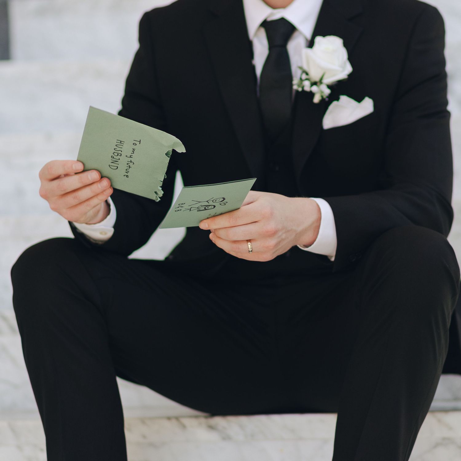 A person in a black suit holds a small green book while sitting on white steps during a formal occasion.