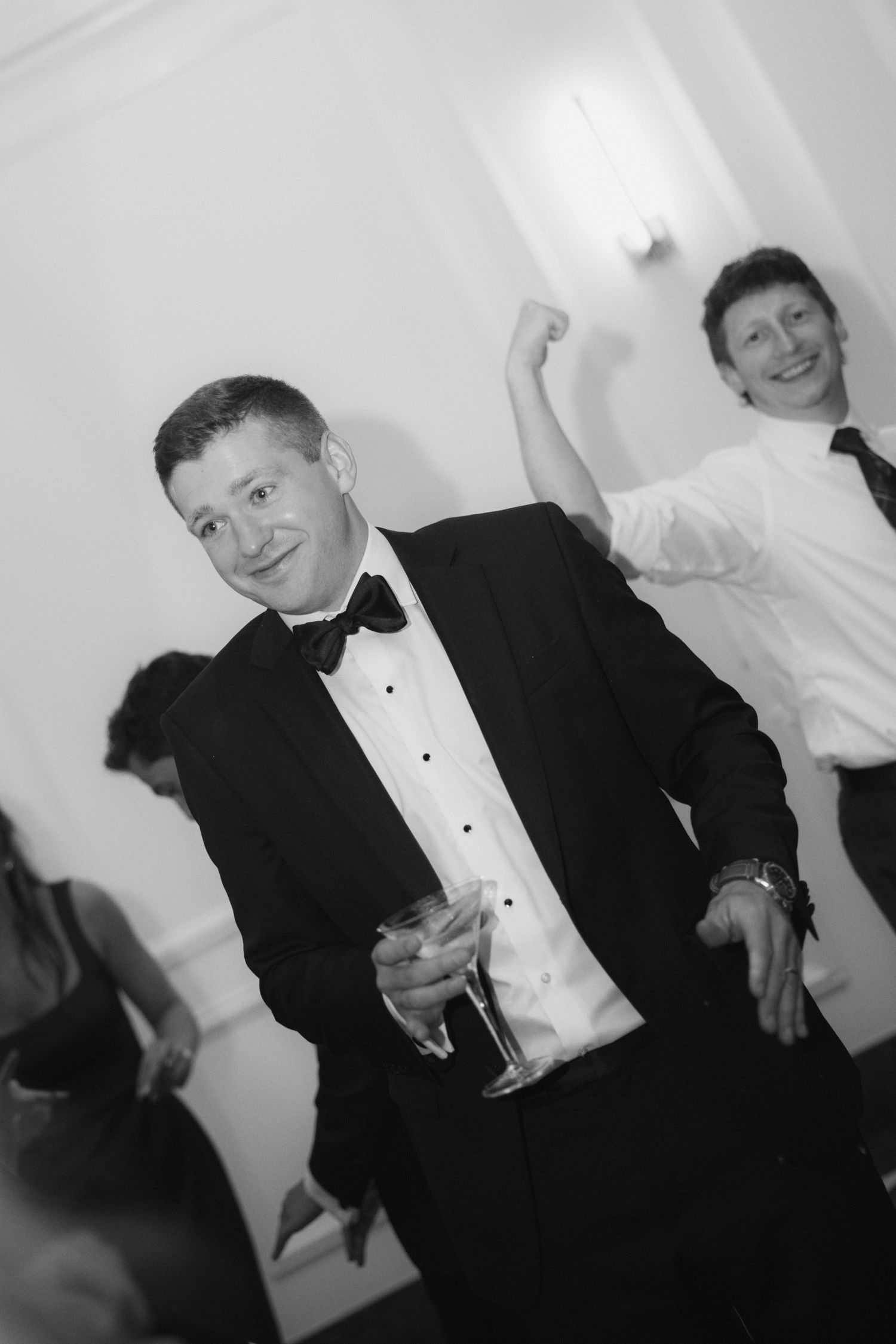 Man in tuxedo with bow tie holds champagne glass while another guest dances behind him at formal event.