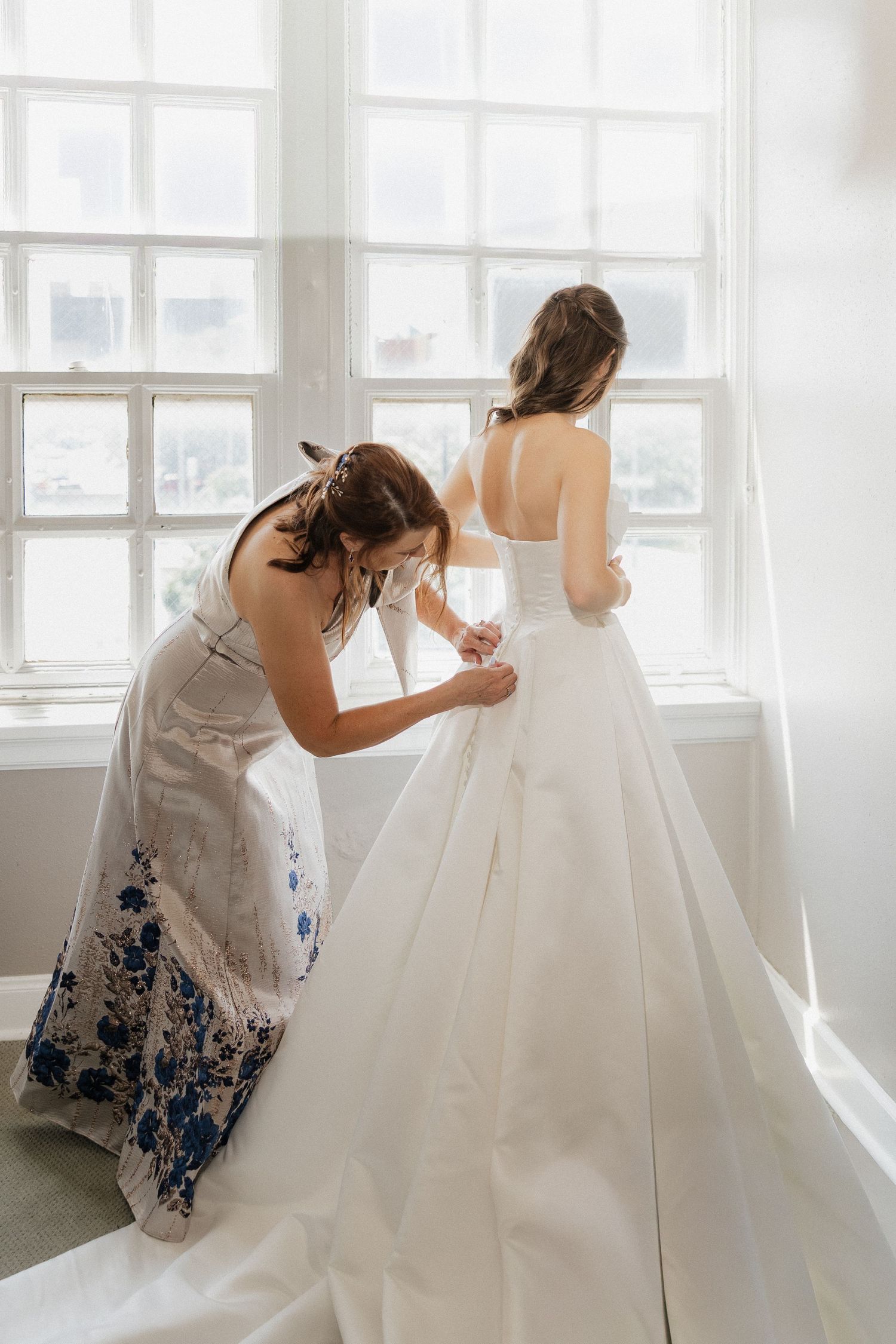 A bridesmaid in a floral dress fastens the back of a bride's white ballgown near a bright window.