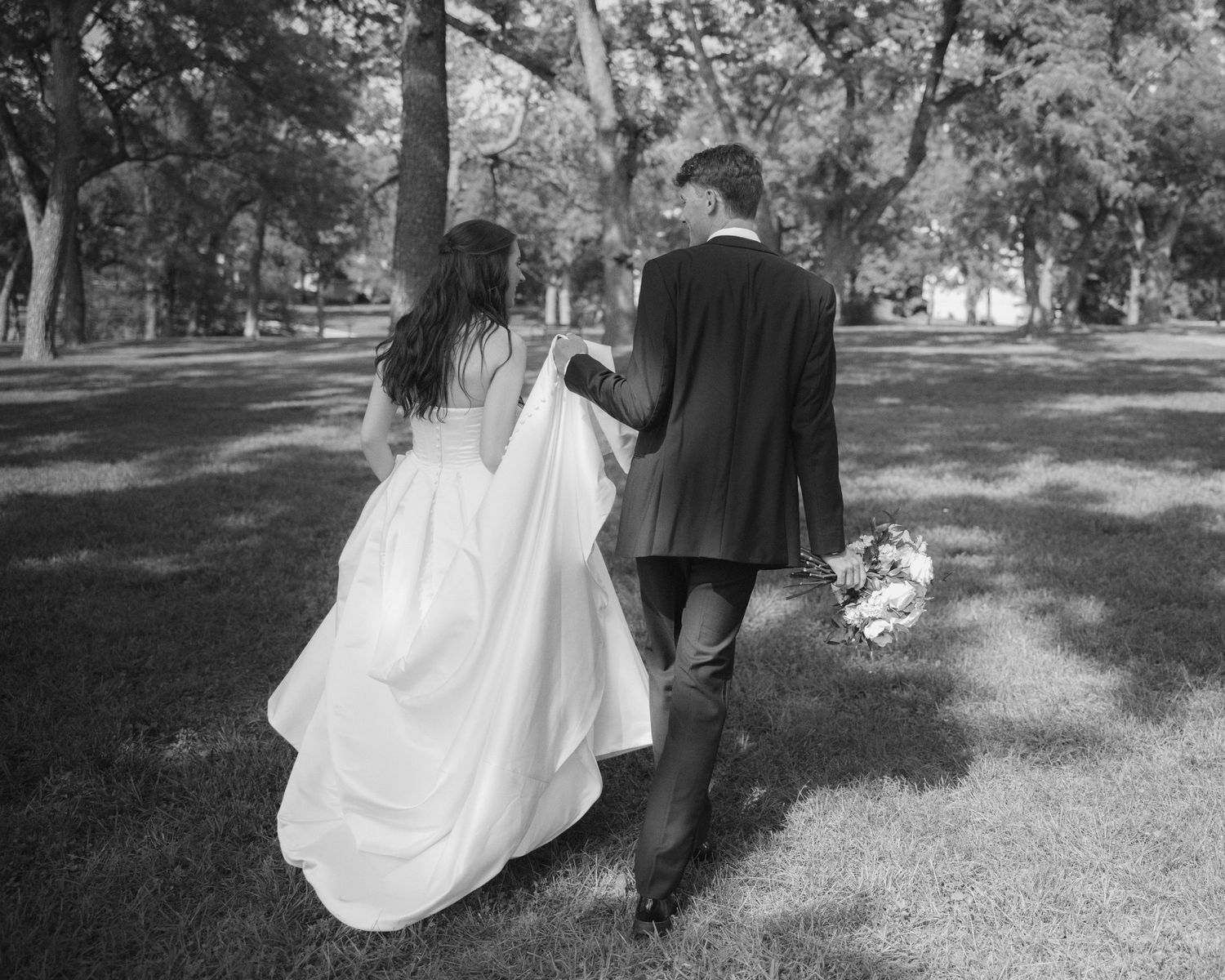 Black and white photo of bride and groom walking hand in hand through a tree-lined park on their wedding day.