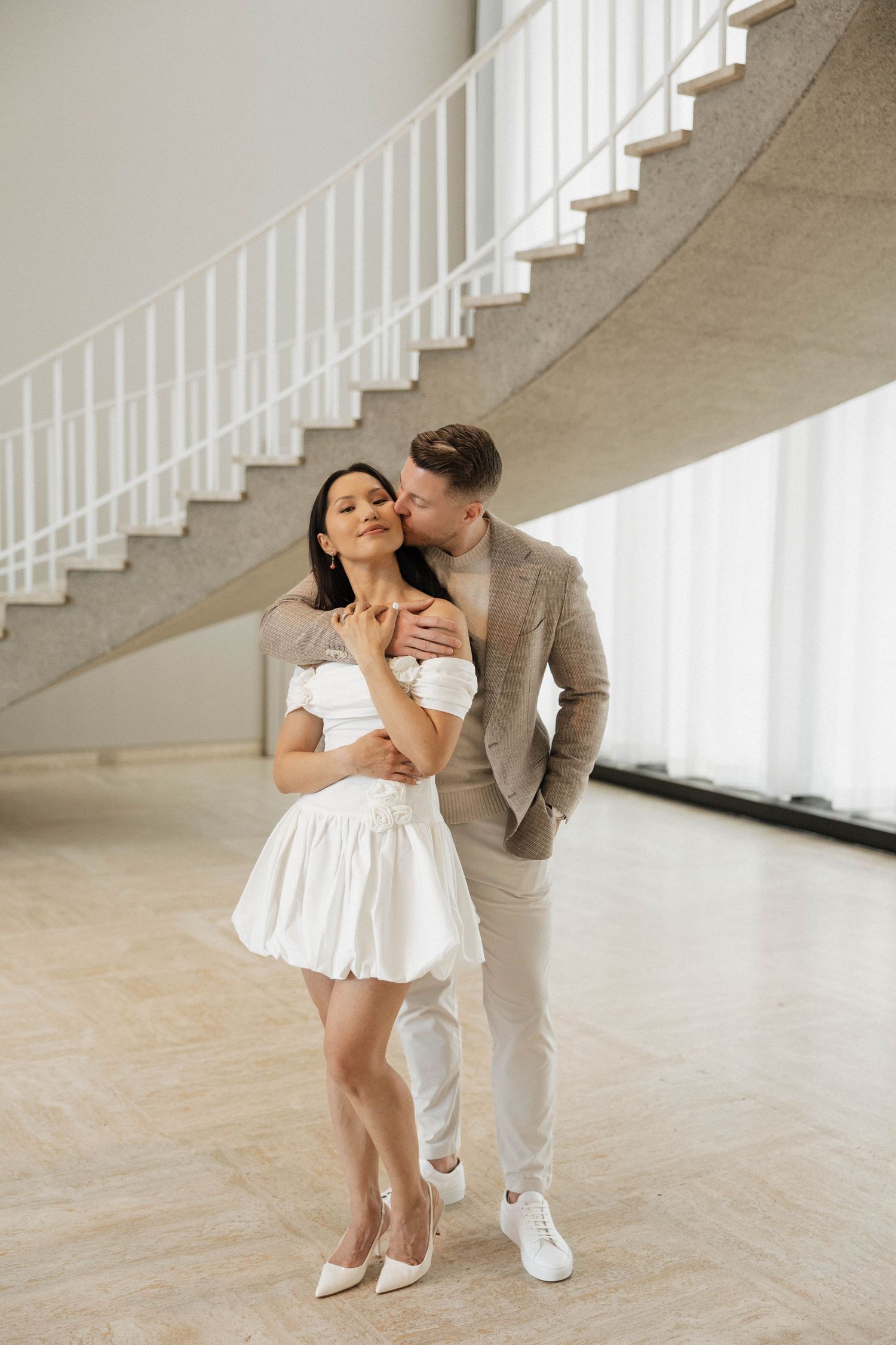 Elegant couple embracing near spiral staircase, woman in white mini dress and man in beige suit in modern interior.