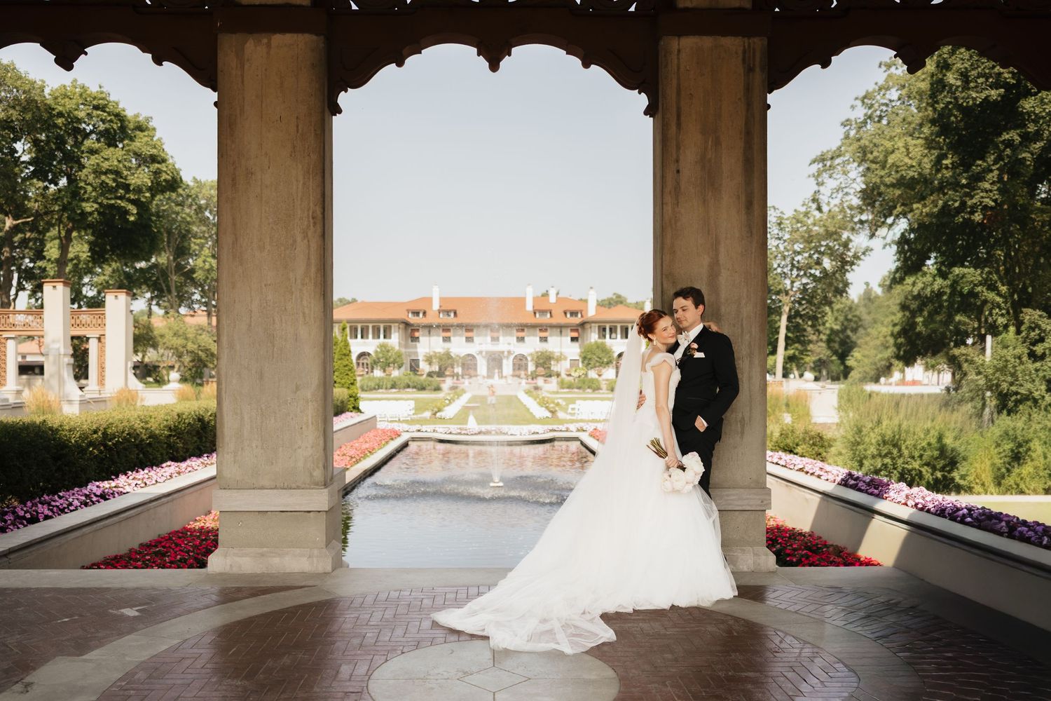 Bride and groom pose under elegant arched colonnade at a formal garden wedding venue with fountain backdrop.