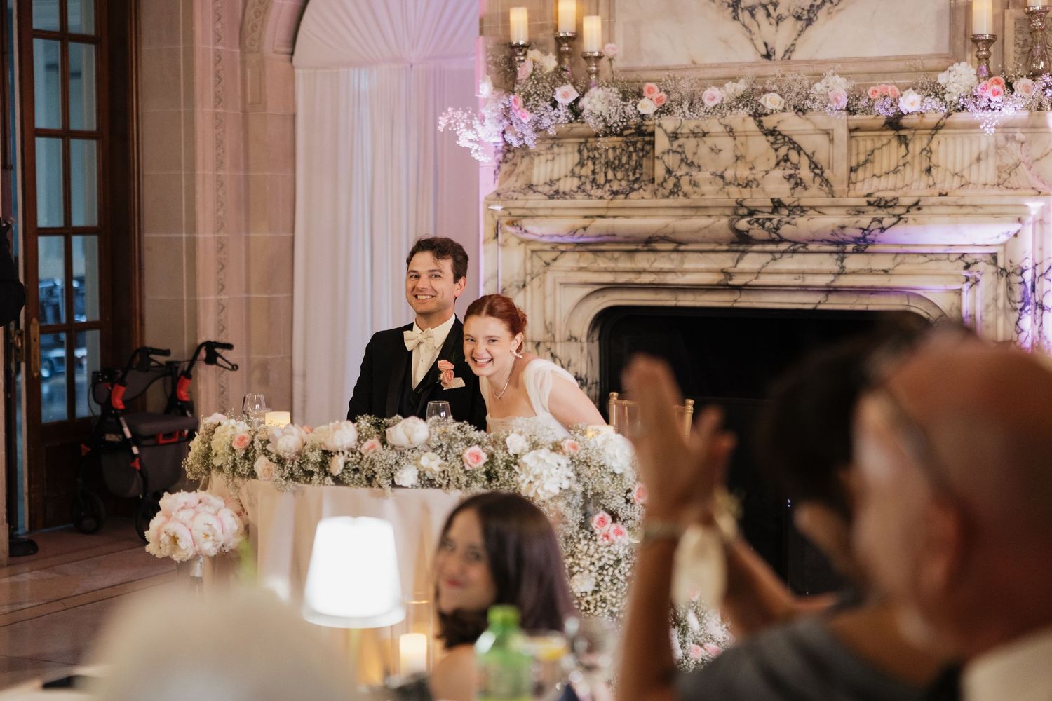 Bride and groom smiling at sweetheart table with floral arrangements at elegant wedding reception.