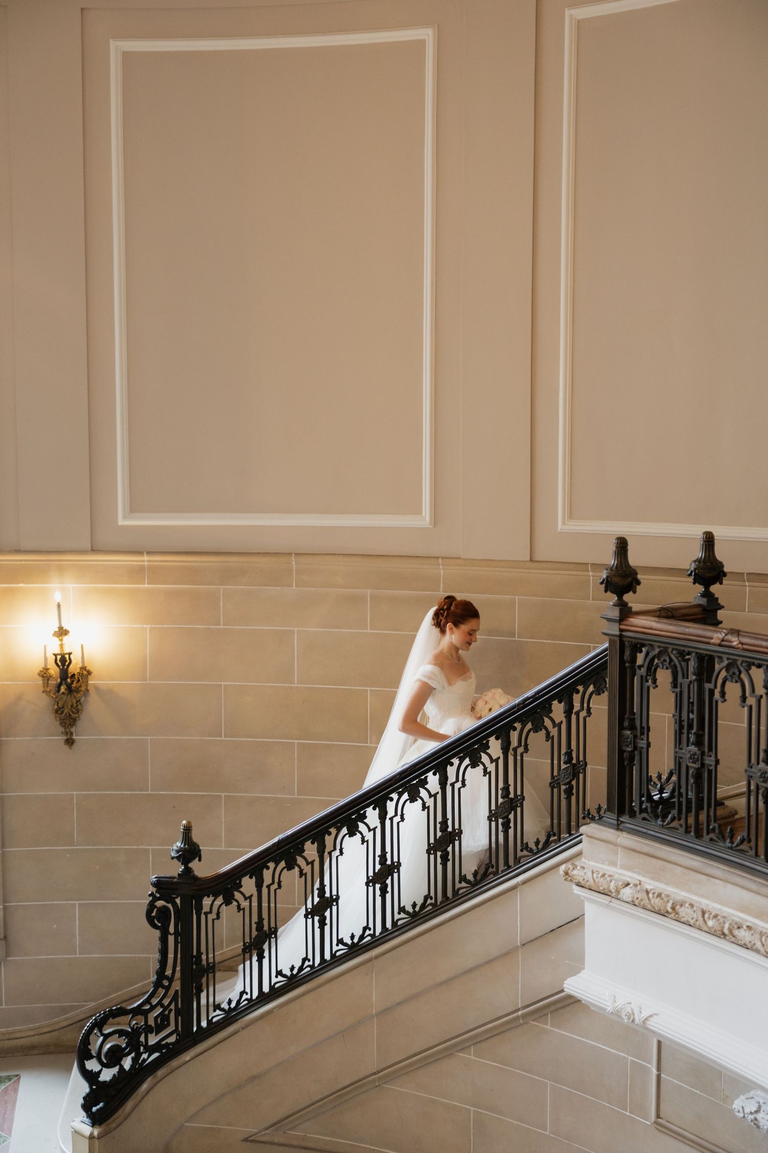 Bride in white gown and veil ascending elegant staircase with ornate black iron railing in a classic venue.