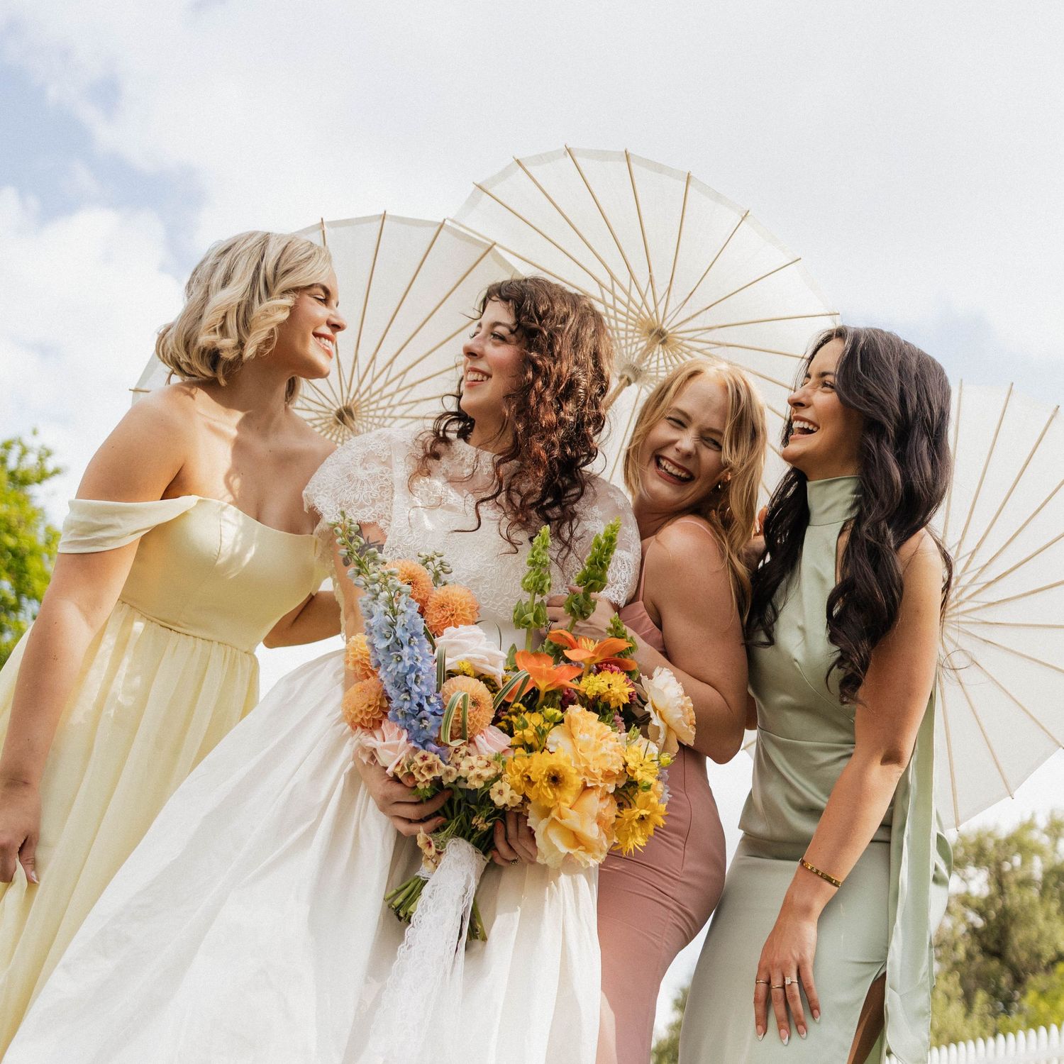Bride holding colorful bouquet laughing with three bridesmaids holding white parasols outdoors.
