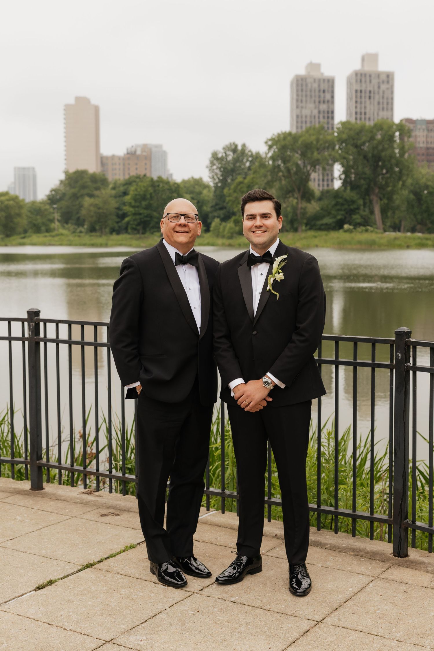 Two men in black tuxedos pose by a lakeside railing with a city skyline backdrop at a wedding.