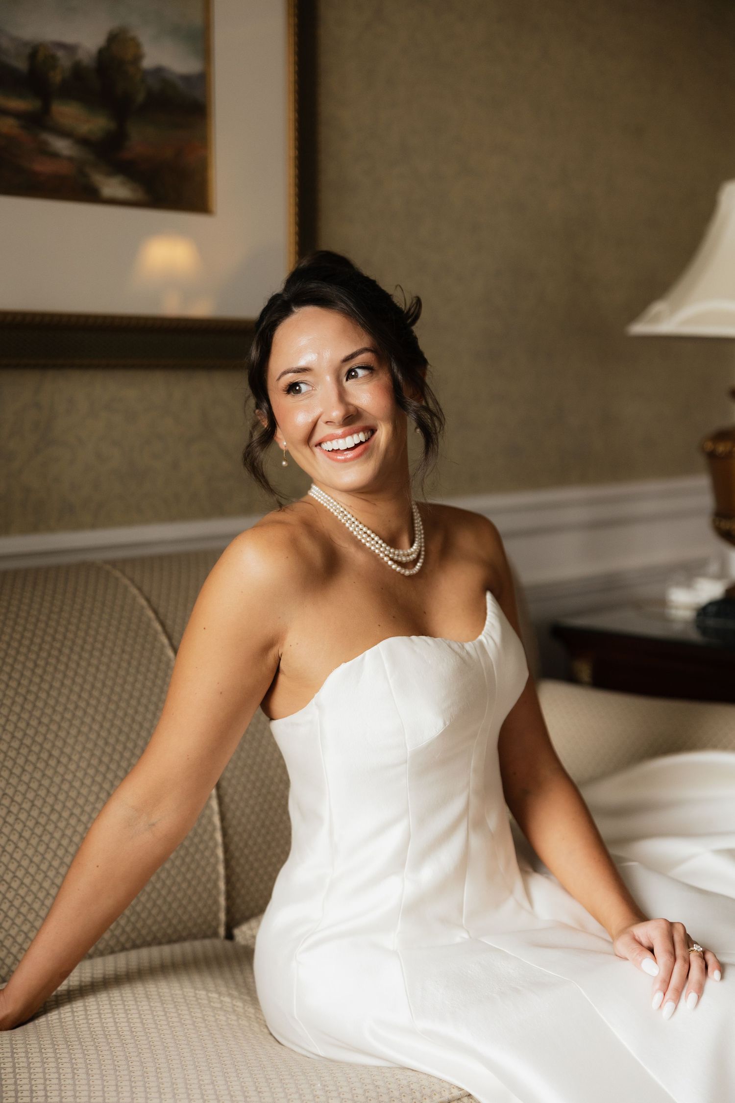 Smiling bride in strapless white gown and pearl necklace sitting elegantly on a cream sofa in a warmly lit room.