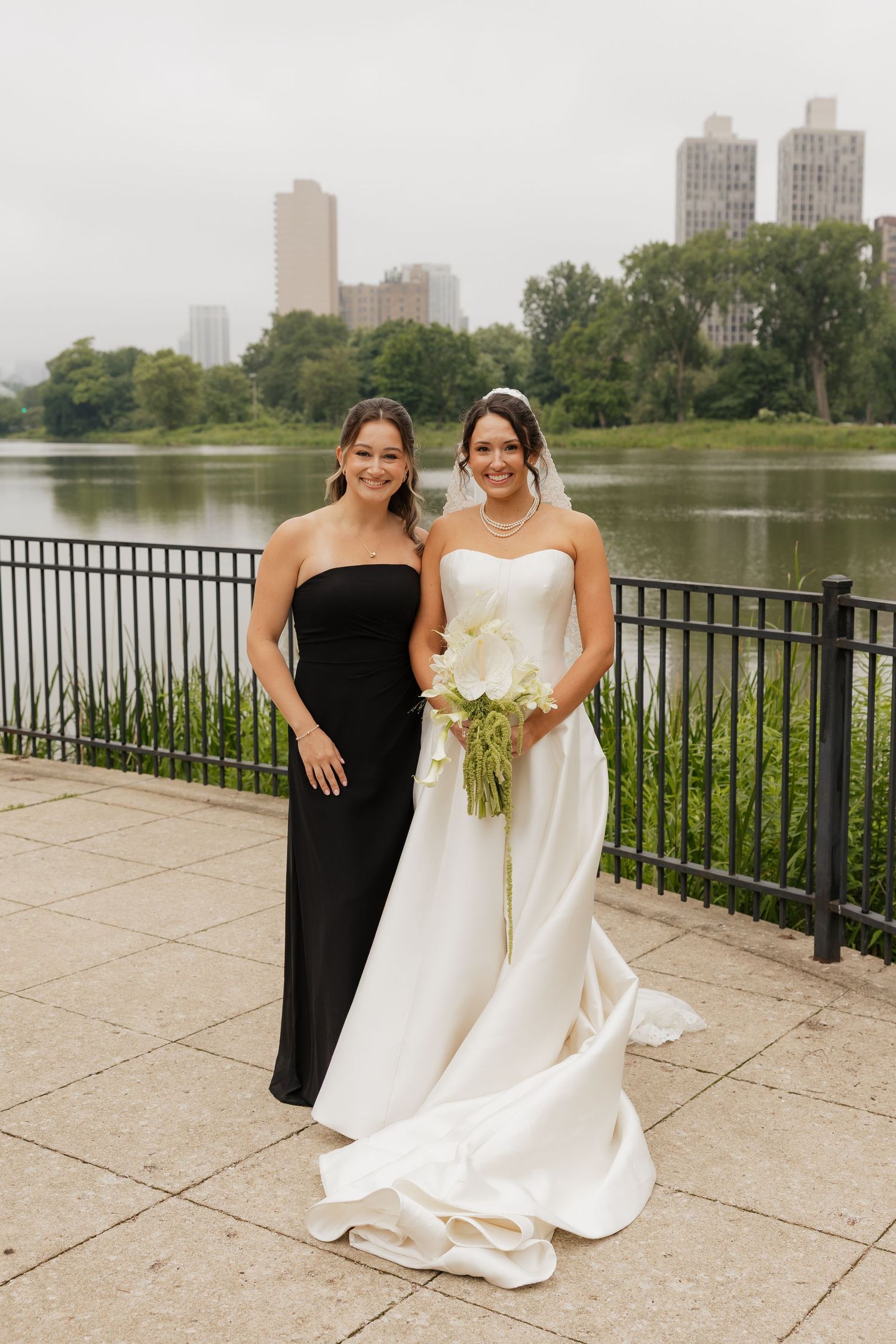 Bride in white gown with bouquet and bridesmaid in black dress posing by a lake in Central Park, NYC.