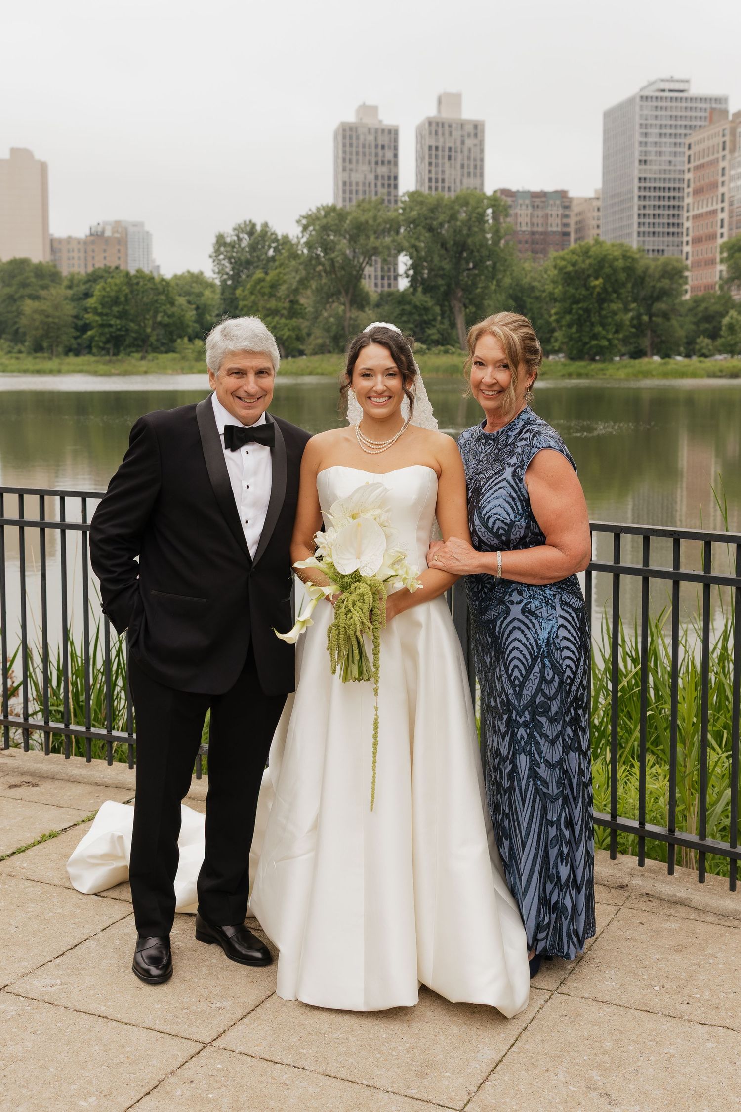 Bride in white gown poses with parents near Central Park lake, man in tuxedo and woman in blue dress.