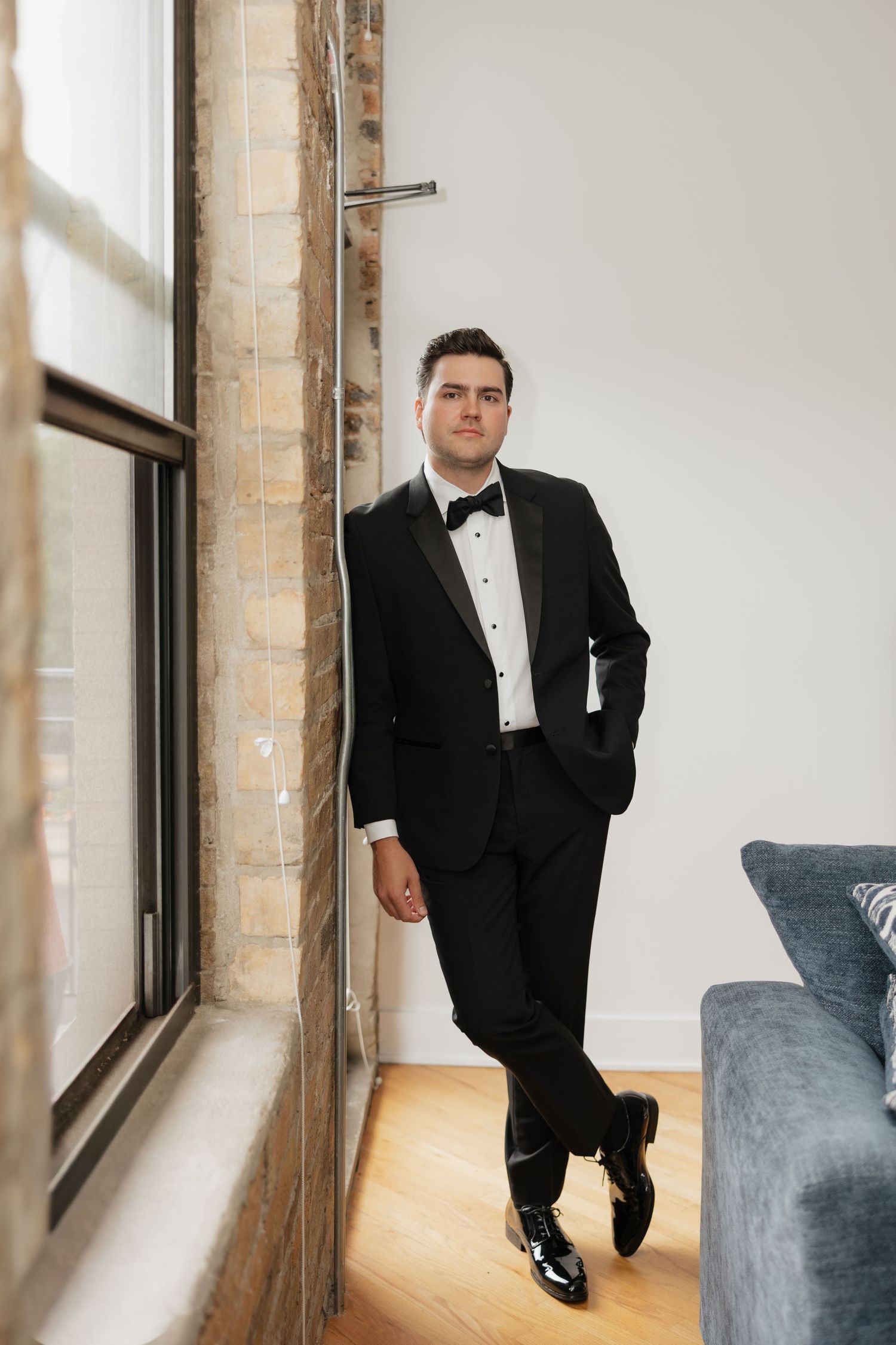 A groom in a black tuxedo with bow tie leans against a brick pillar near a window in a bright modern room.