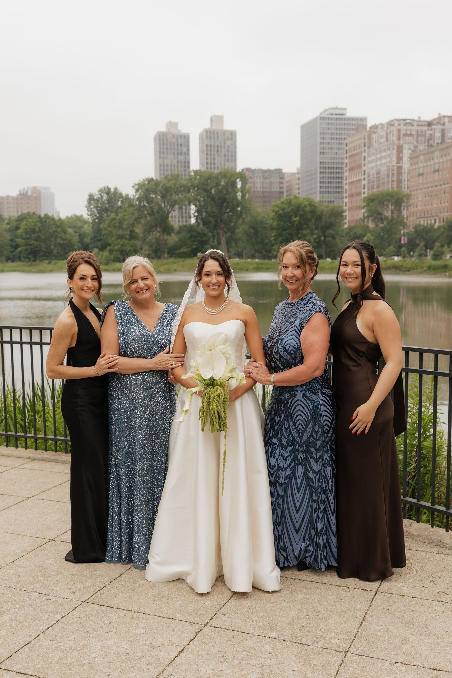 Bride in white gown poses with four bridesmaids in blue and black dresses by a lake with city skyline backdrop.