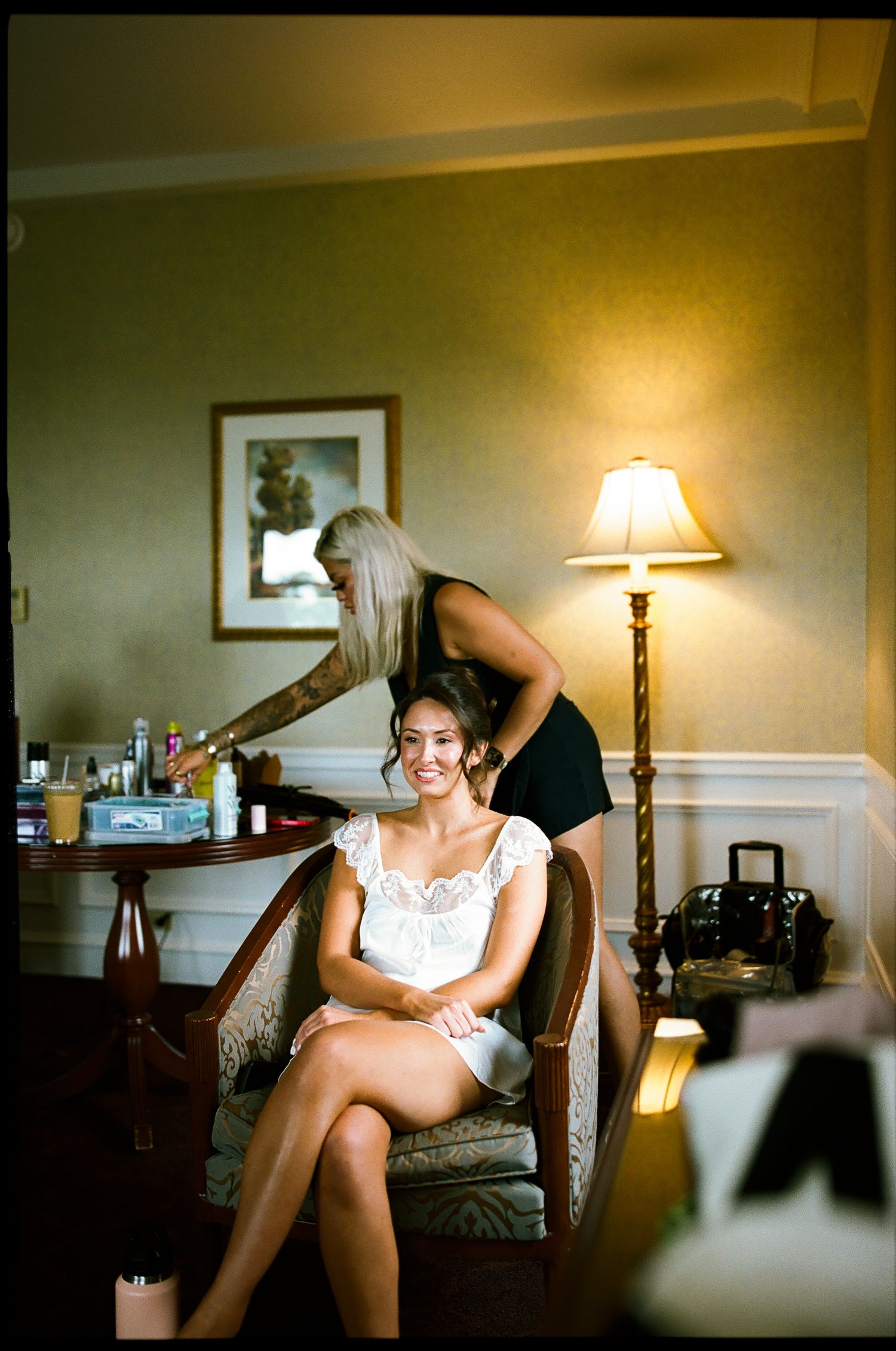 Bride smiling in chair while hairstylist works in warmly lit hotel room before wedding.