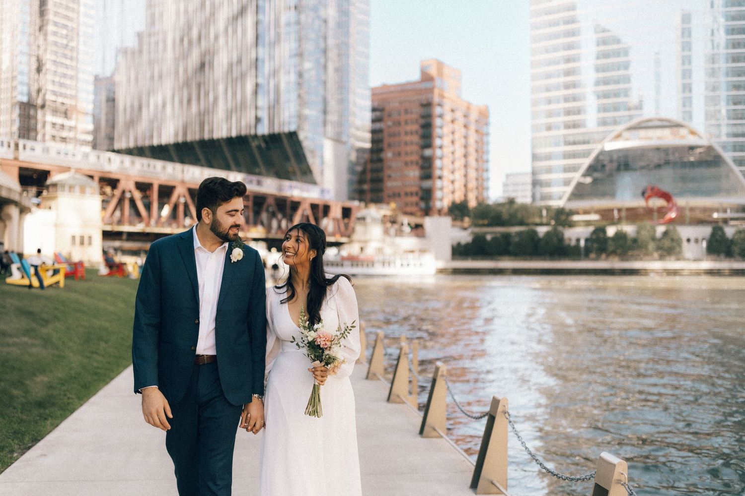 Couple stands together at a waterfront plaza with city buildings and fountains in the background.