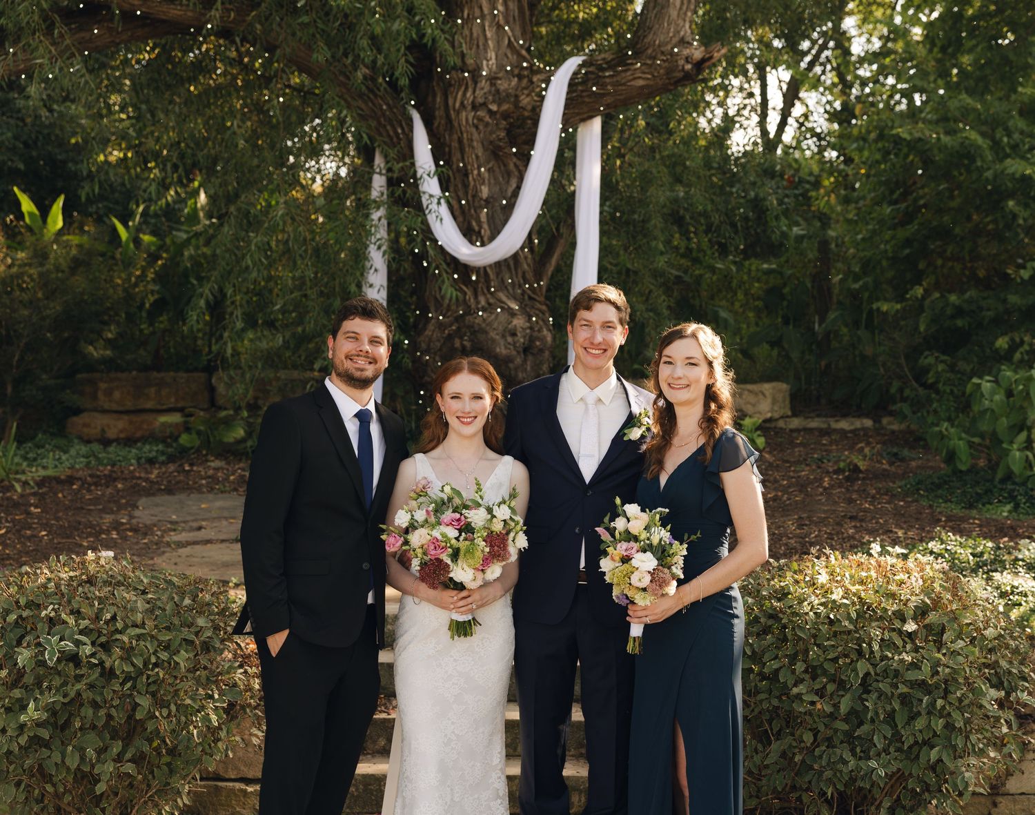 Wedding party in formal attire standing together in an outdoor garden setting.