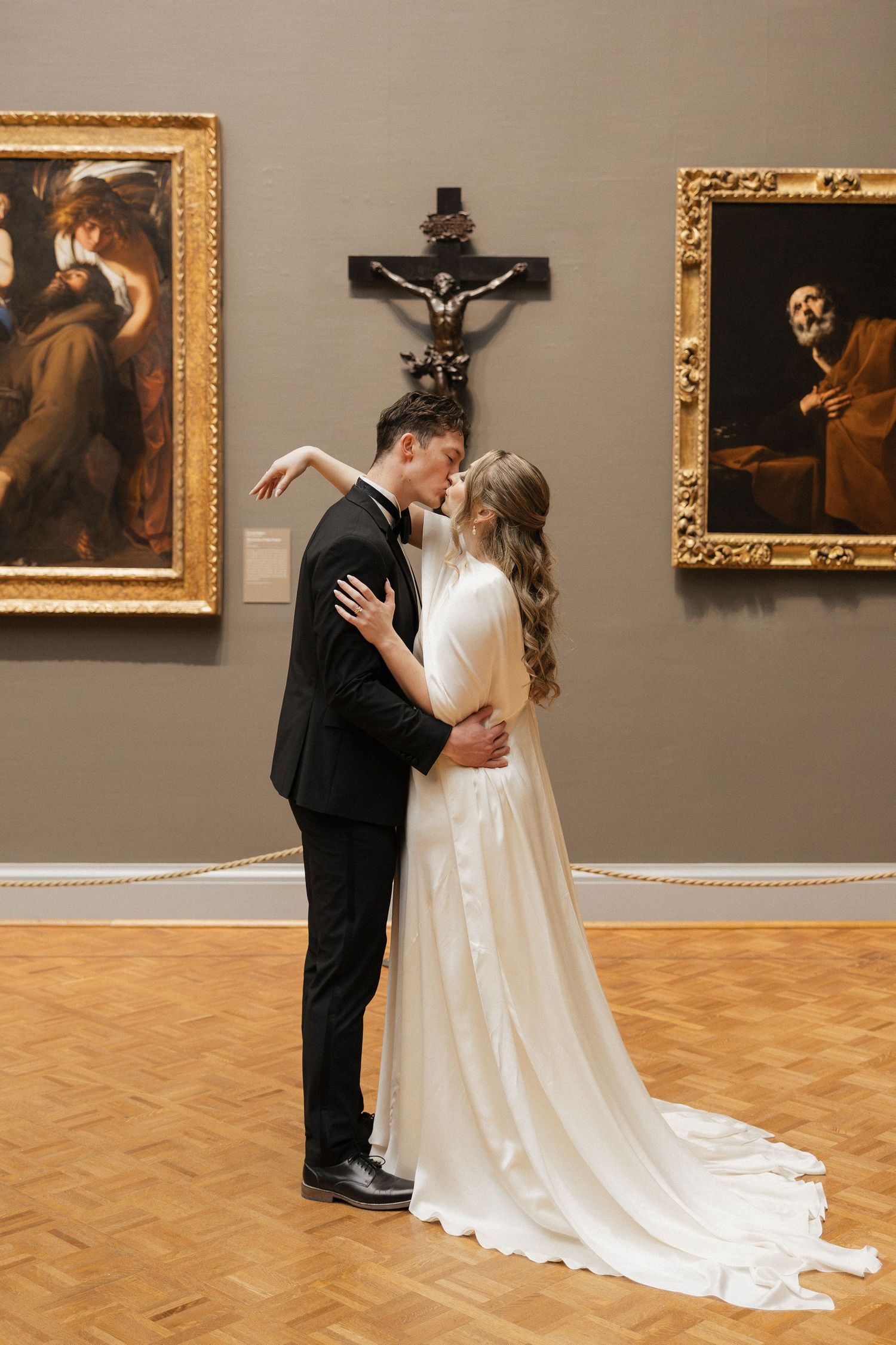 A romantic wedding portrait of a couple embracing in an art gallery with religious paintings and a crucifix.