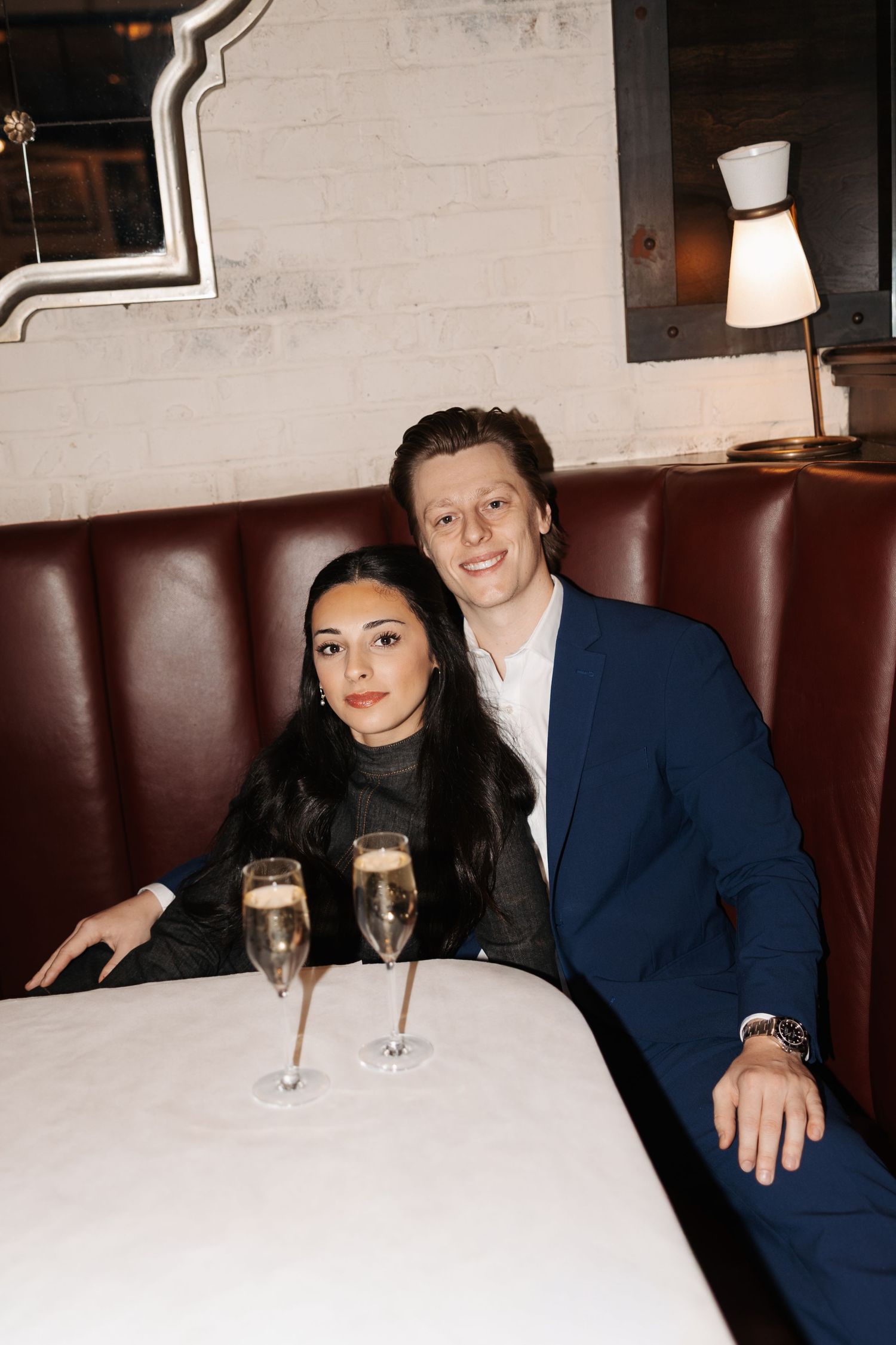 A couple sits at a restaurant booth with champagne glasses on the white tablecloth in a dimly lit setting.