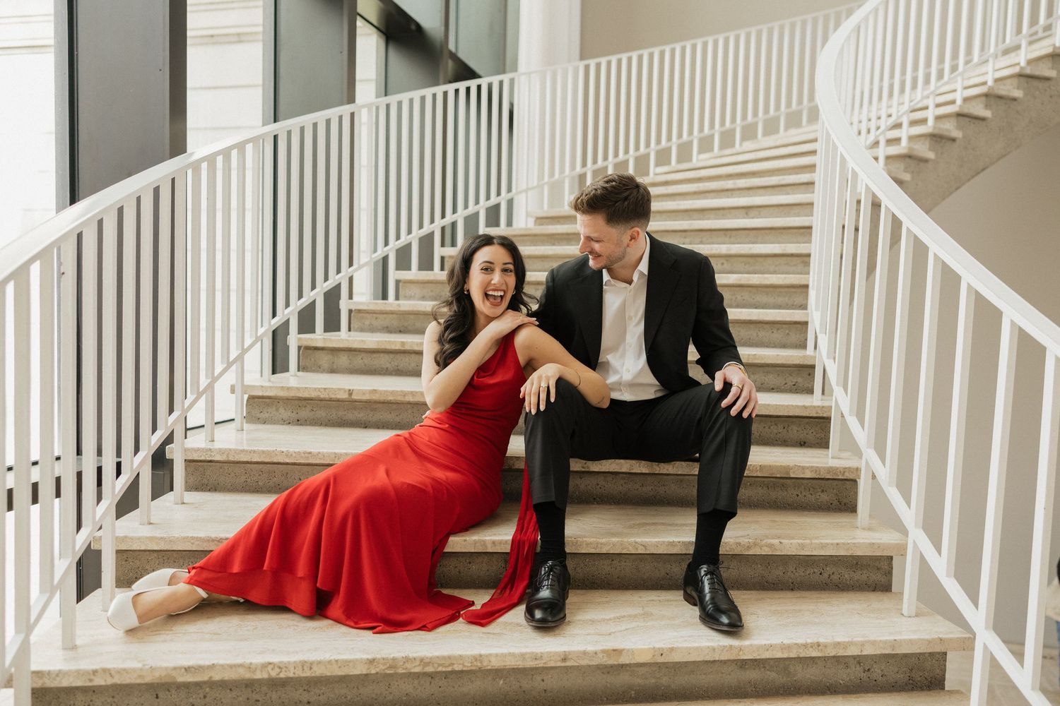 Couple in formal attire poses romantically on white marble staircase, with woman wearing flowing red dress.