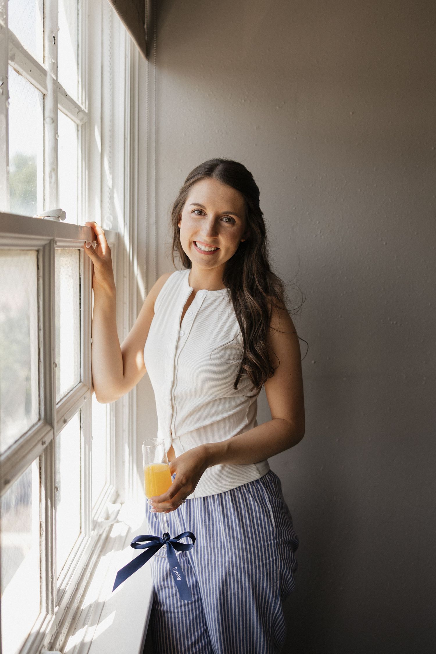 Young woman in white top and striped pajama pants smiling by window holding glass of orange juice.