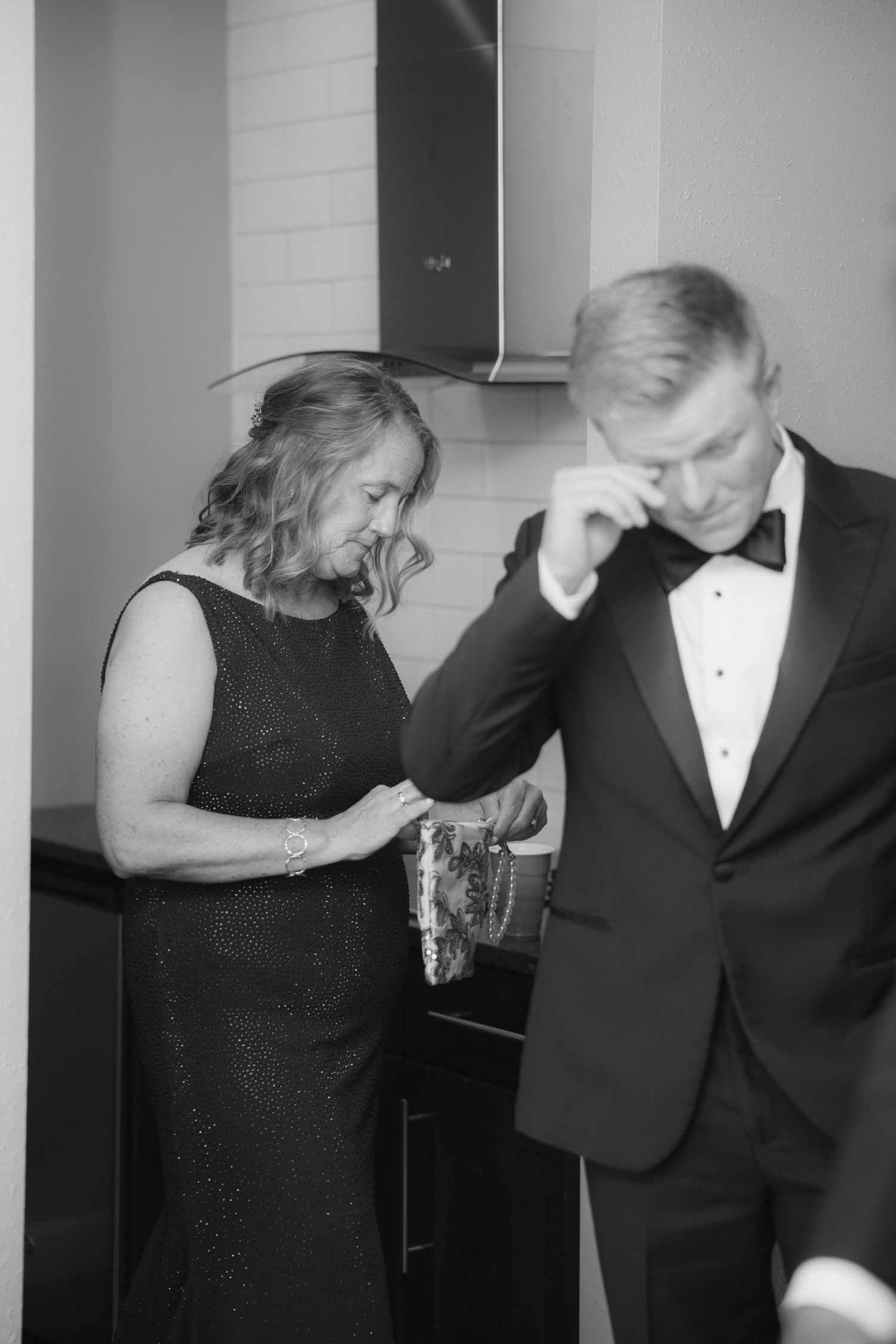 Black and white photo of a groom wiping tears while his mother adjusts his tuxedo jacket before the wedding ceremony.