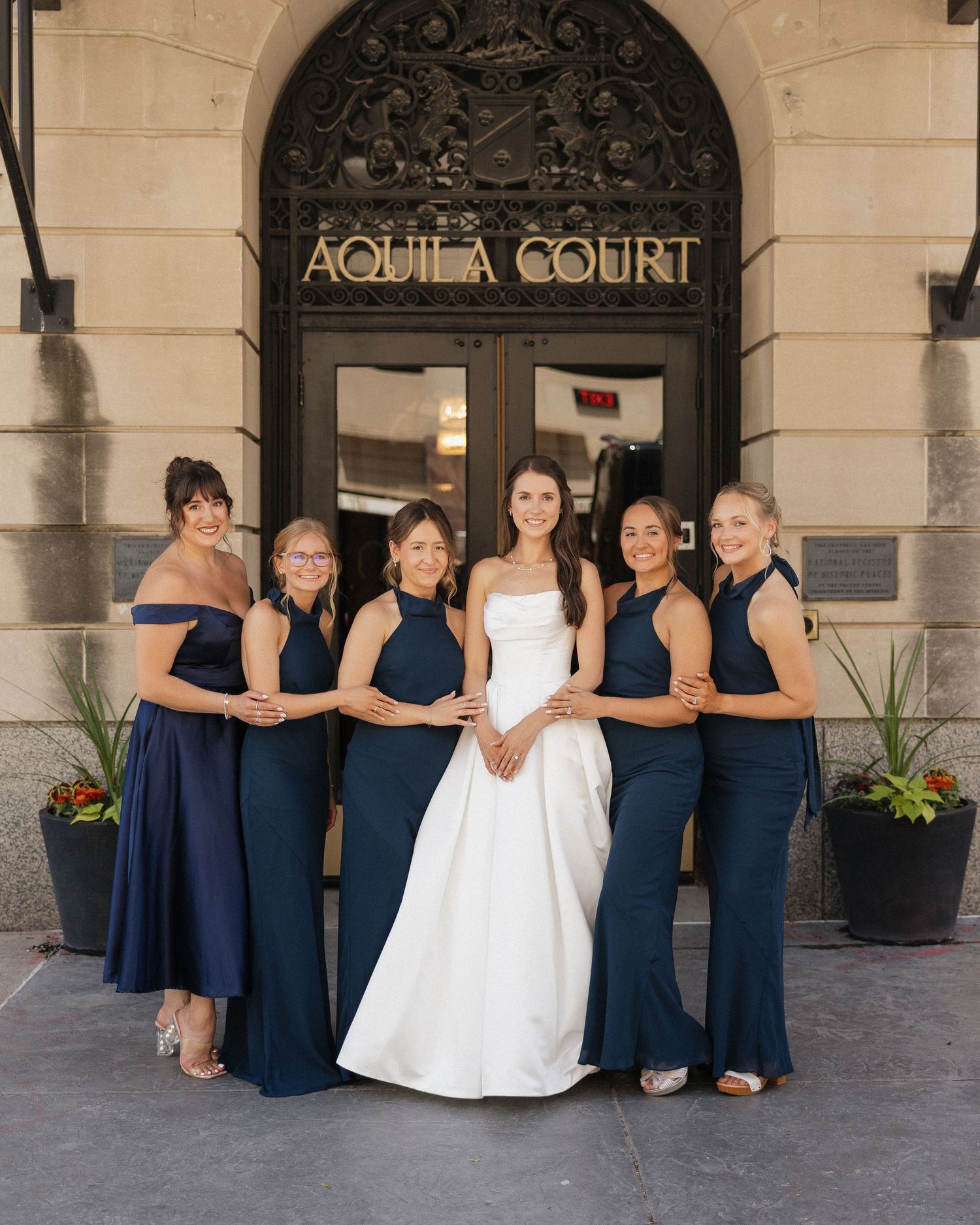 Bride in white gown with five bridesmaids in navy dresses posing outside Aquila Court building entrance.