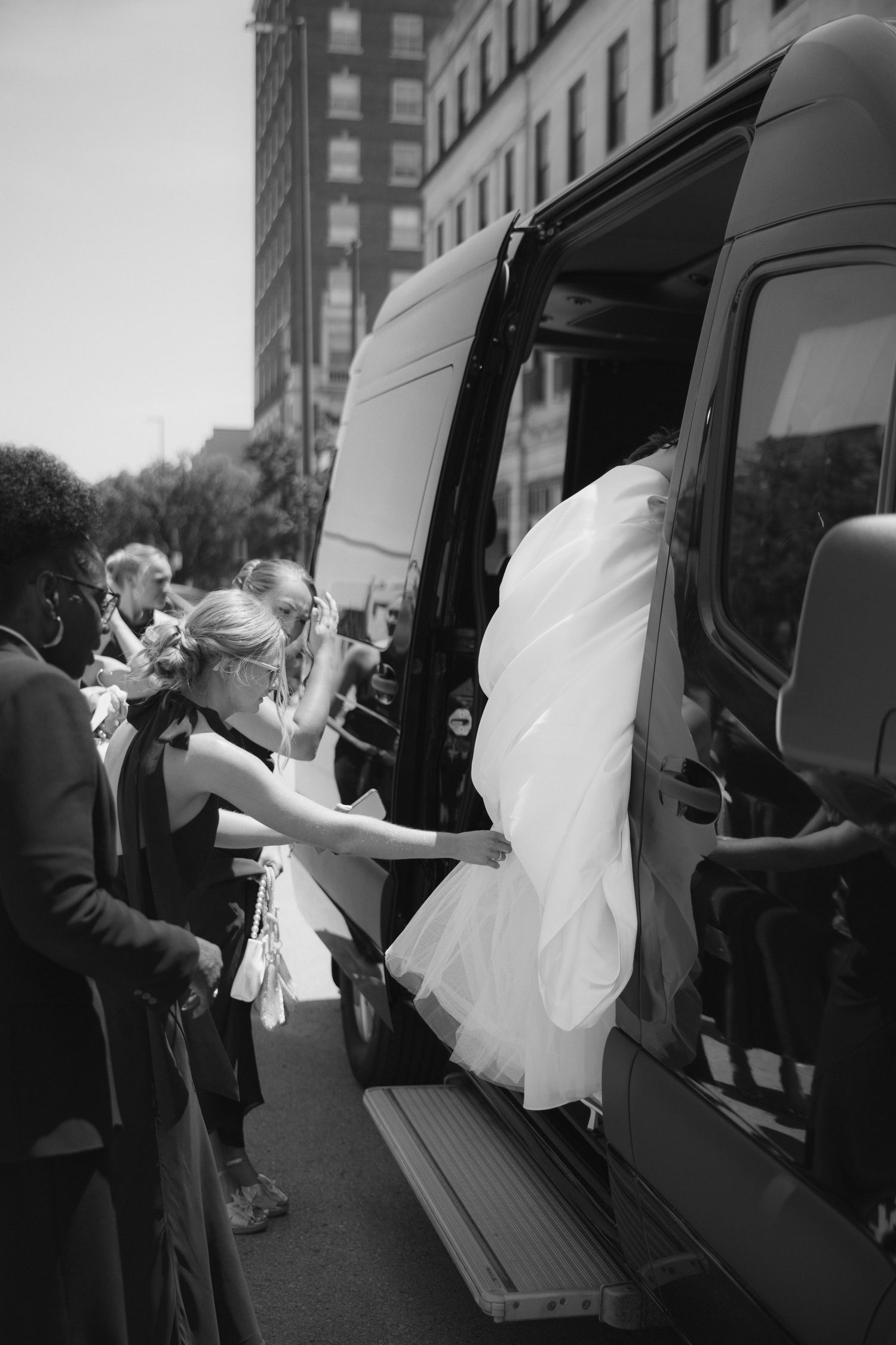 Bride in white gown boarding a black van assisted by bridesmaid, wedding party gathered outside urban building.