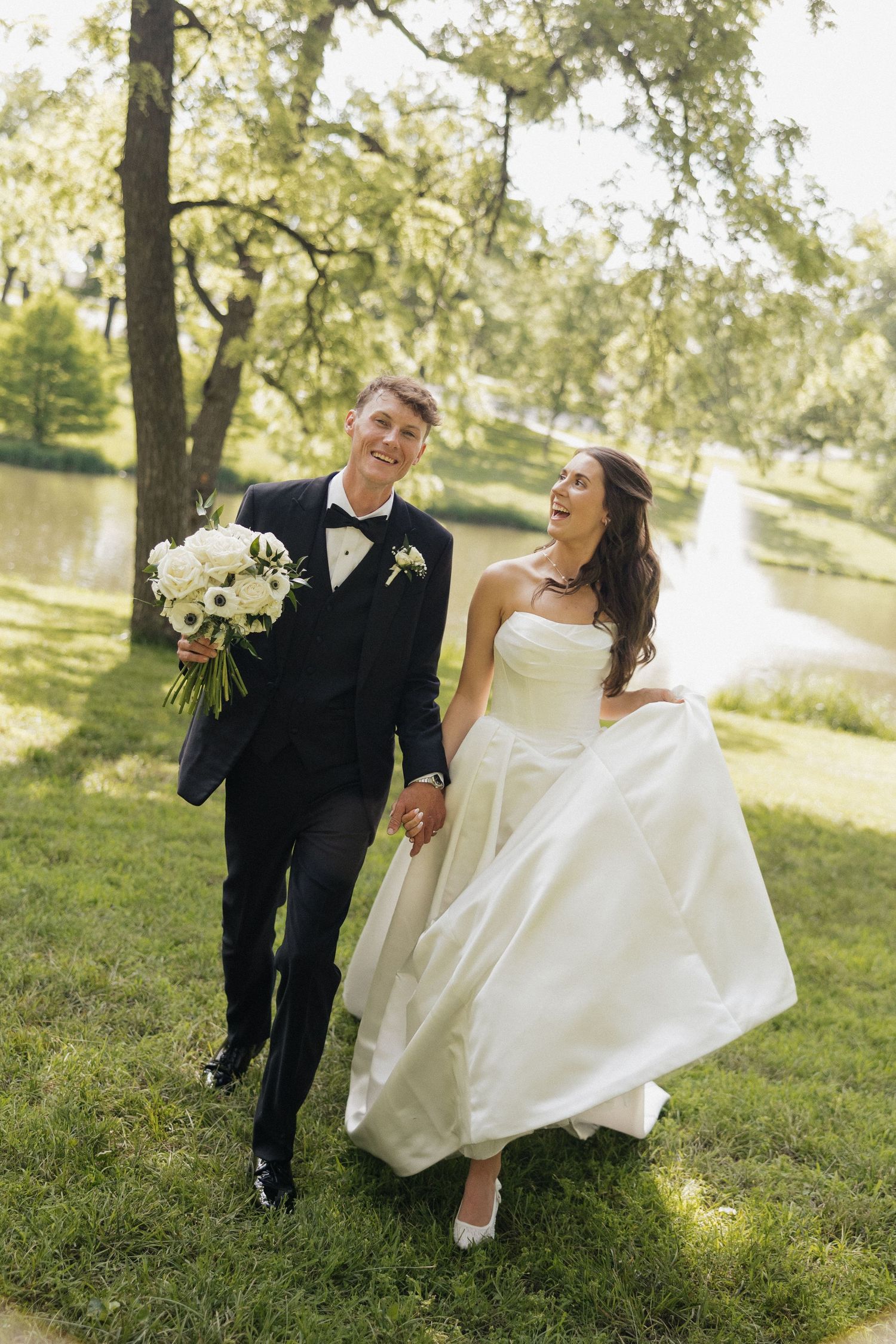 Happy newlyweds walk hand-in-hand through a lush green park, bride in white gown holding dress, groom in black tux with bouquet.