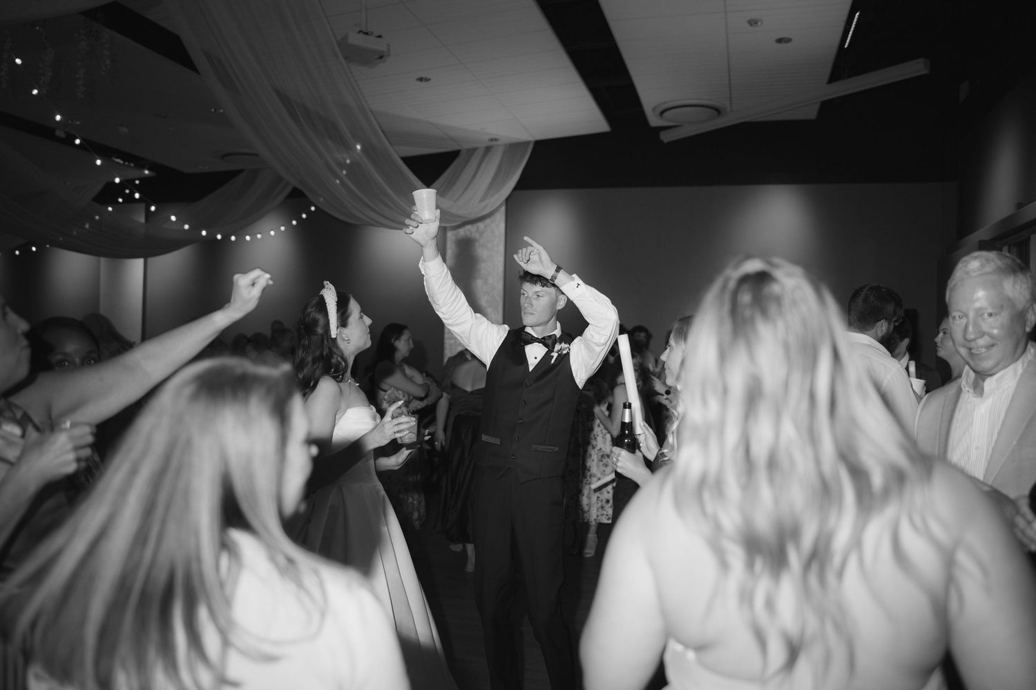 Black and white photo of a groom dancing joyfully with guests at a lively wedding reception celebration.
