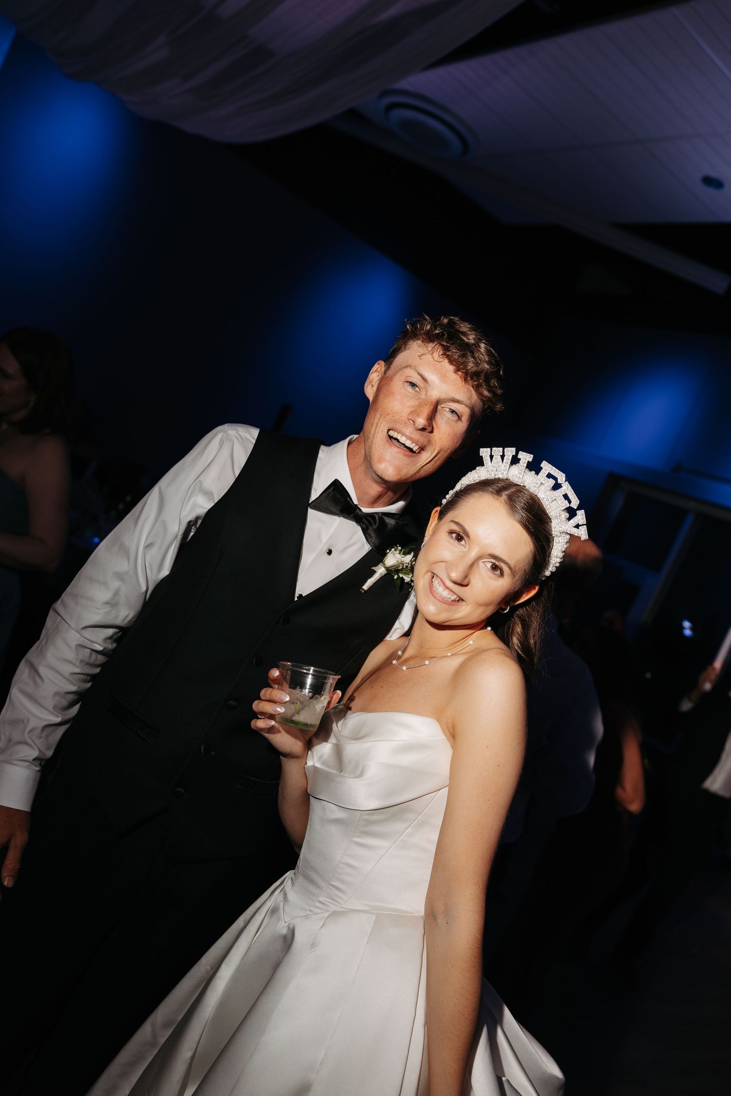 A smiling bride in a white strapless gown and tiara poses with a groom in a black tuxedo at their wedding reception.