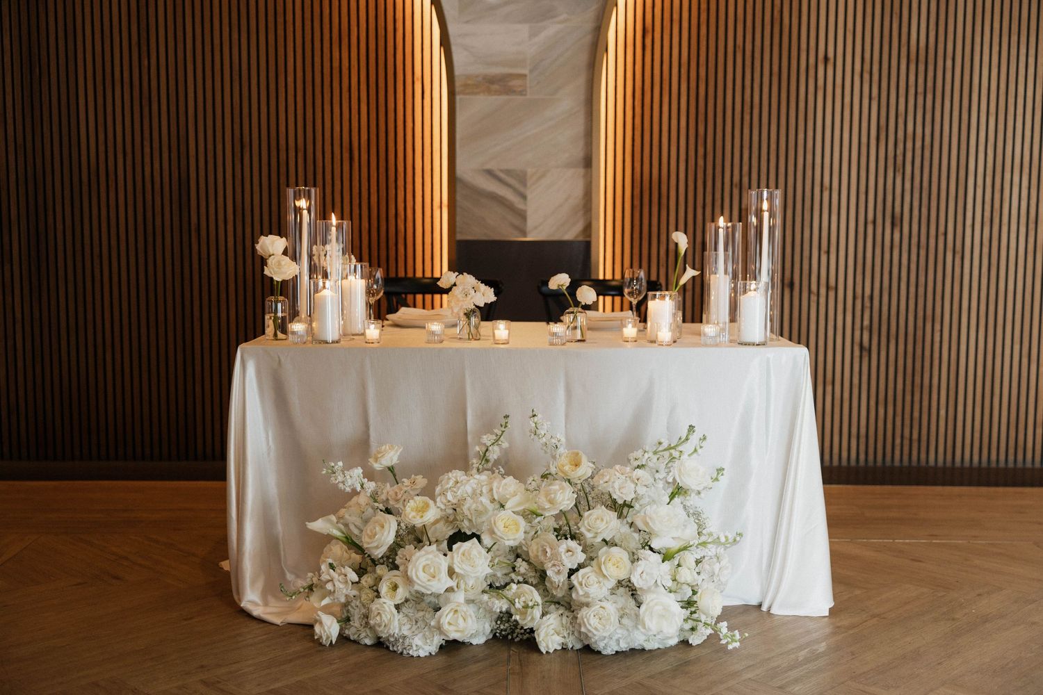 Elegant wedding sweetheart table with white floral arrangements, candles, and warm wood backdrop.