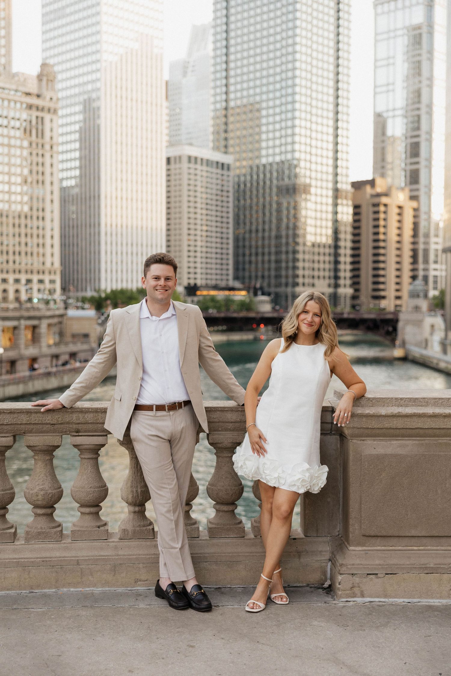 Couple in elegant attire poses on Chicago Riverwalk bridge with stunning city skyline backdrop.