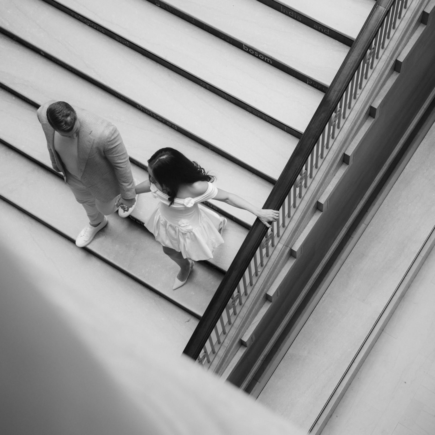Aerial black and white photo of a couple holding hands walking on striped steps of a modern building.