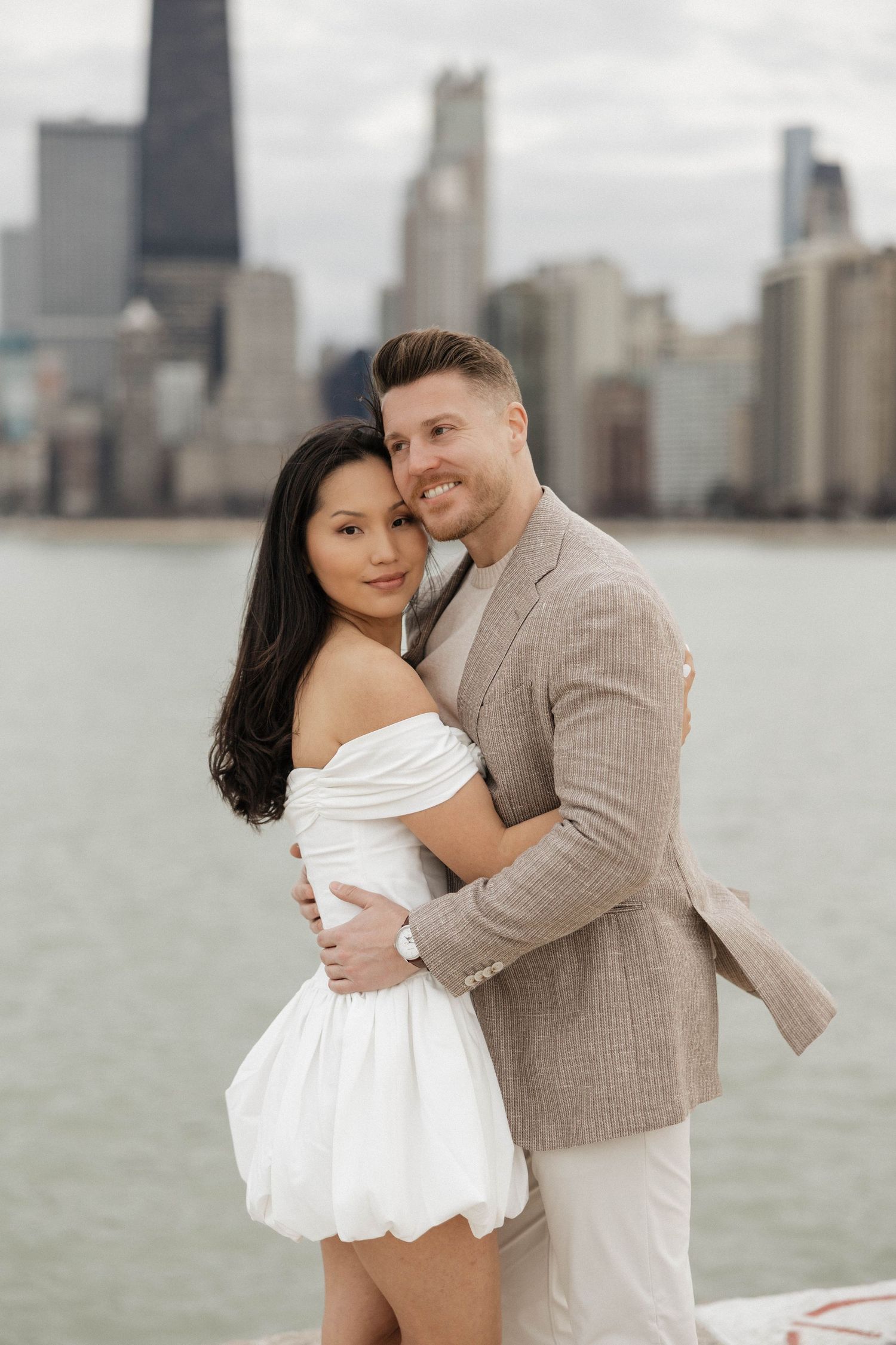 Couple embracing and smiling by Chicago skyline waterfront, woman in white off-shoulder dress, man in tan blazer.