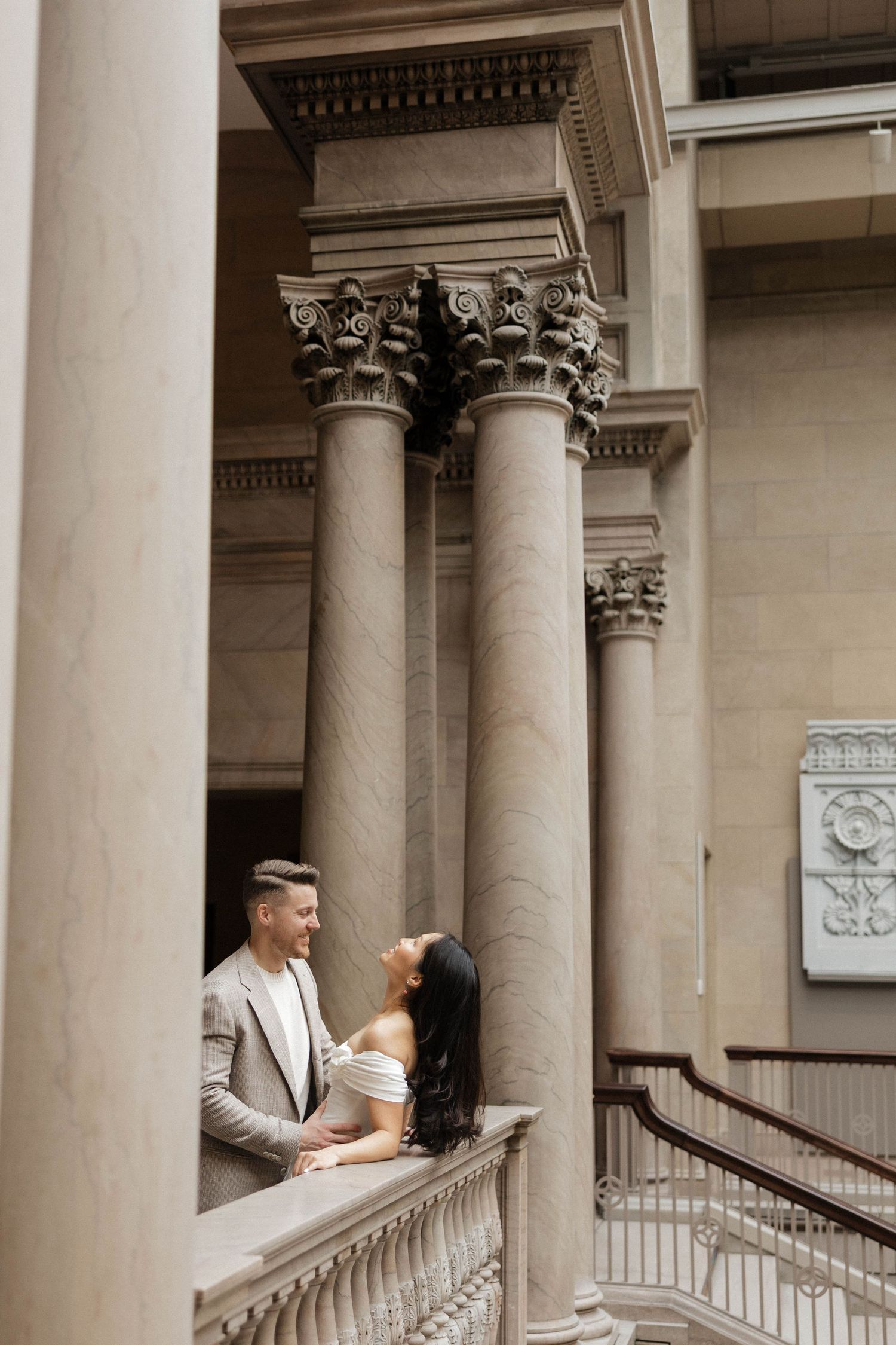 Couple posing romantically on a grand staircase with ornate Corinthian columns in an elegant classical building.