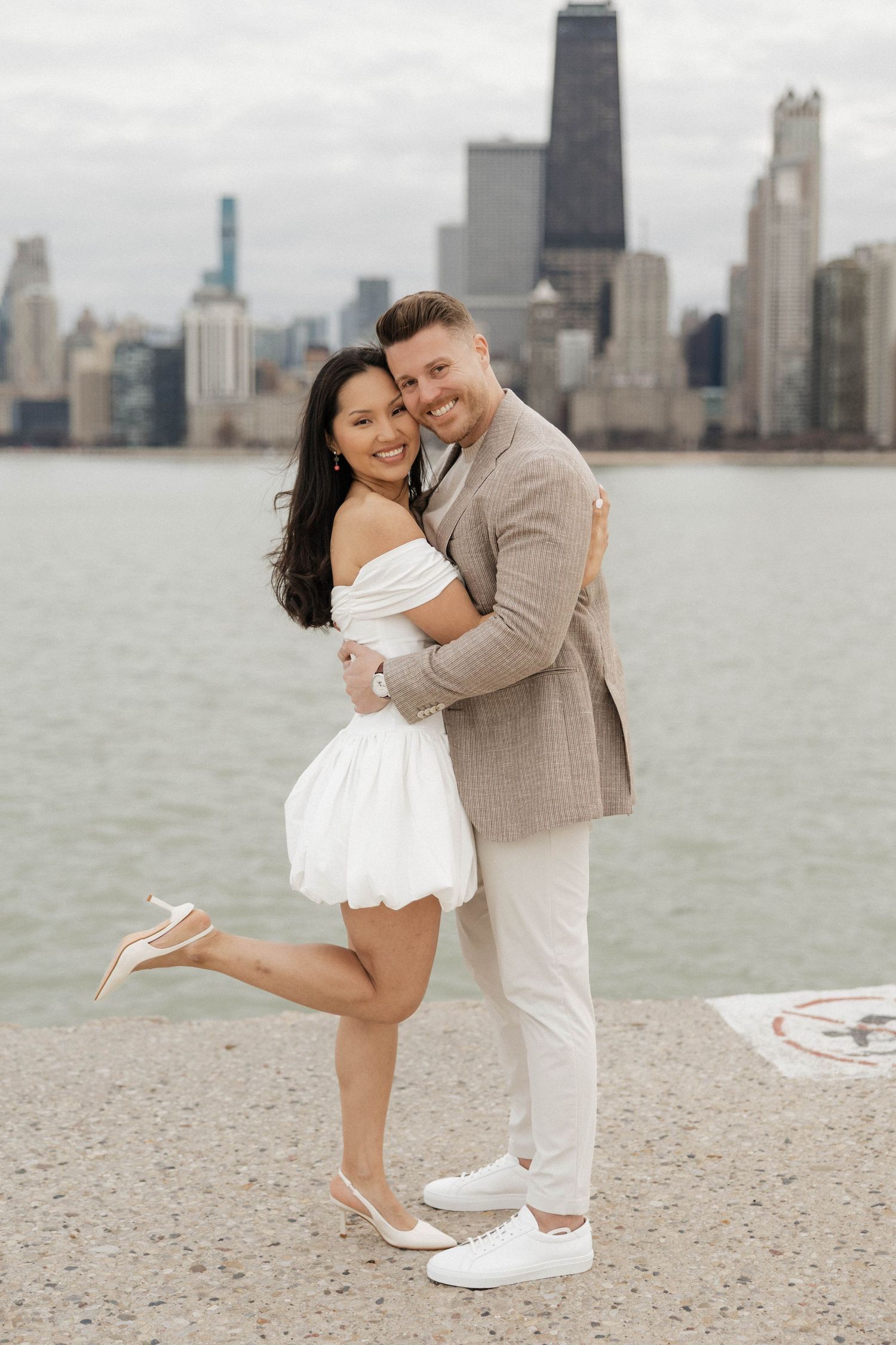 Couple embracing and smiling by Chicago skyline waterfront, woman in white off-shoulder dress, man in beige blazer.